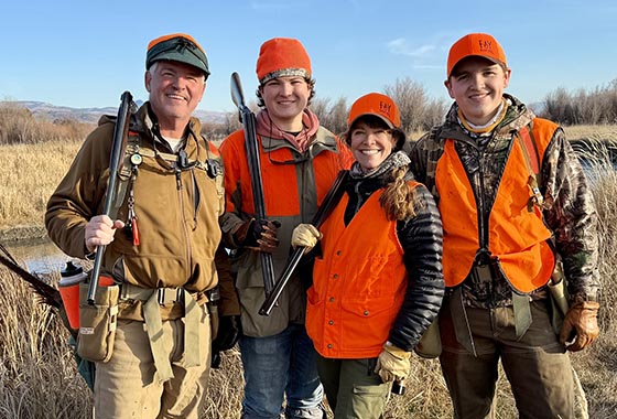 Four people outdoors in a grassy wetland, wearing orange safety vests and hats, holding shotguns and posing for a group photo.