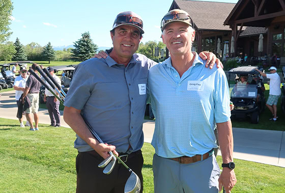 Two men standing arm-in-arm on a golf course, one holding golf clubs, with a clubhouse and spectators in the background. Name tags are visible on their shirts.