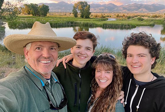 Selfie of a man wearing a wide-brimmed hat with three smiling teens by a river, mountains in the distance at sunset/spring light.