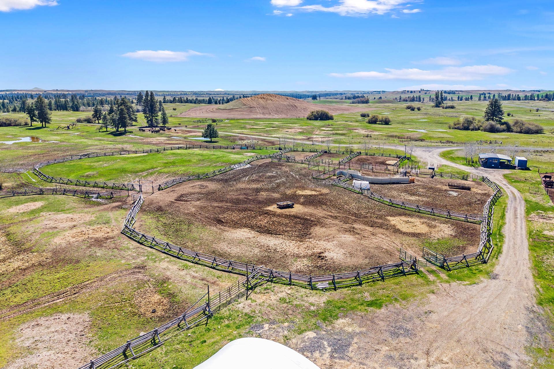 Aerial view of a cattle ranch with circular paddocks and dirt arenas under a blue sky, distant hill on the horizon.