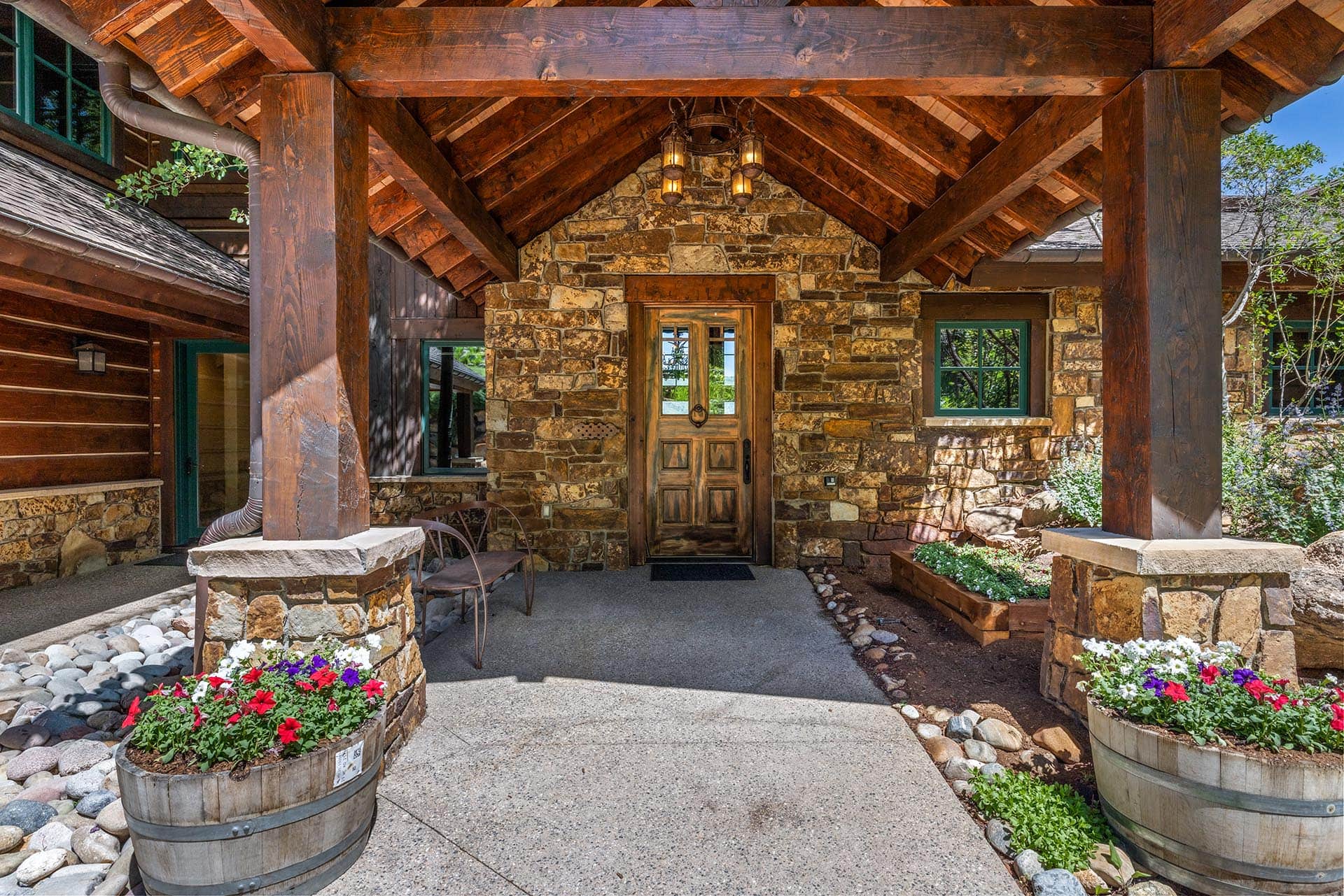 Stone cottage entrance with a timber-framed porch, heavy wooden columns, and a stone façade with flowering barrels nearby.