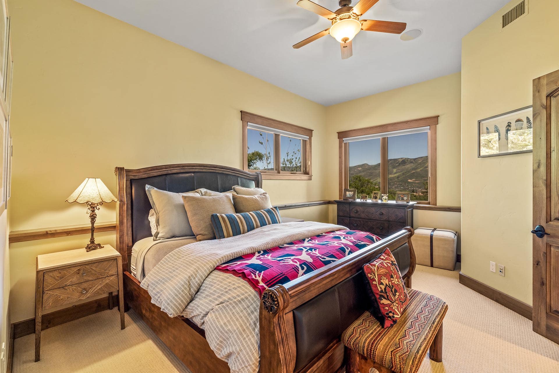 Bedroom with a large wooden bed, yellow walls, and a ceiling fan; windows show mountains outside along with a dresser and lamps.
