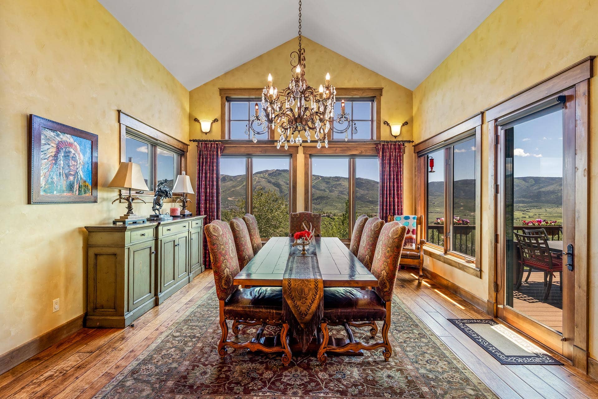 Elegant dining room with a long wooden table, ornate upholstered chairs, and a crystal chandelier, yellow textured walls, and mountains visible through large windows.