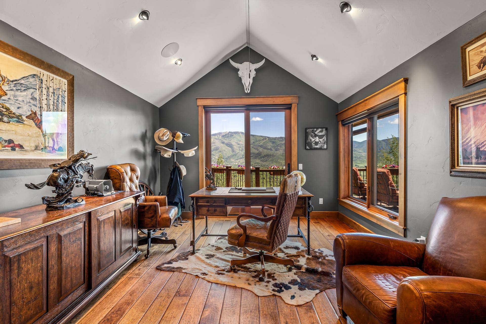 Rustic home office with a wooden desk and leather chairs, cowhide rug, and a mountain view through large windows; bull skull decor above the window.
