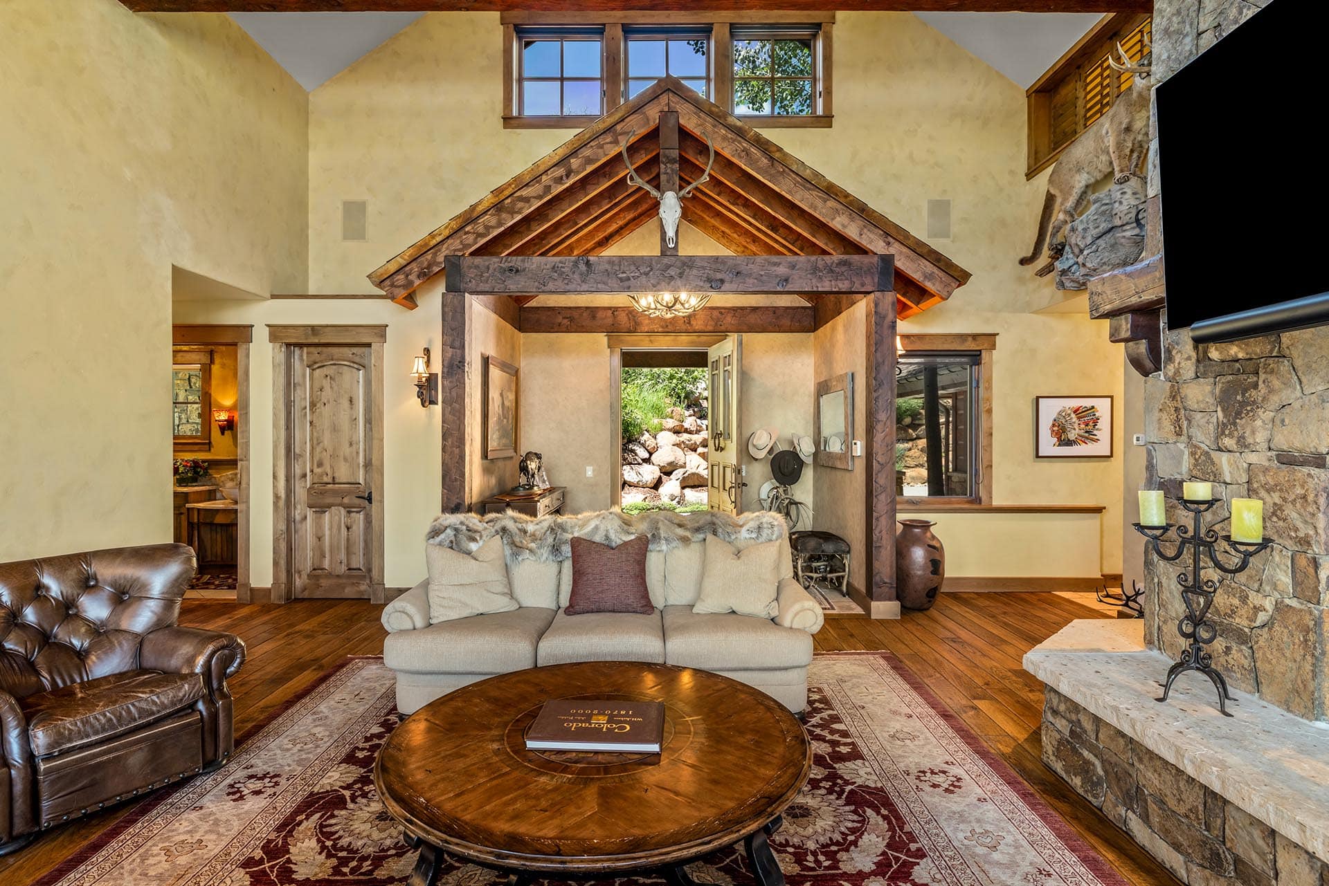 Cozy rustic living room with a stone fireplace, wooden beams, and a large beige sofa facing a round wooden coffee table atop a patterned rug.
