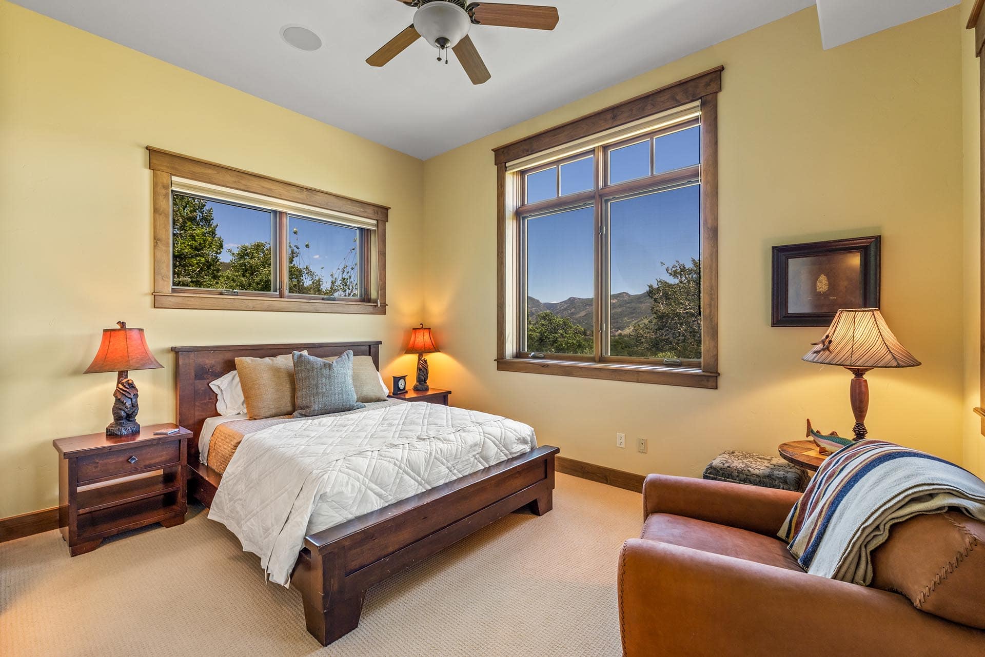 Cozy bedroom with a wooden bed, matching nightstands, and yellow walls; large windowed views of hills outside; two lamps lit.