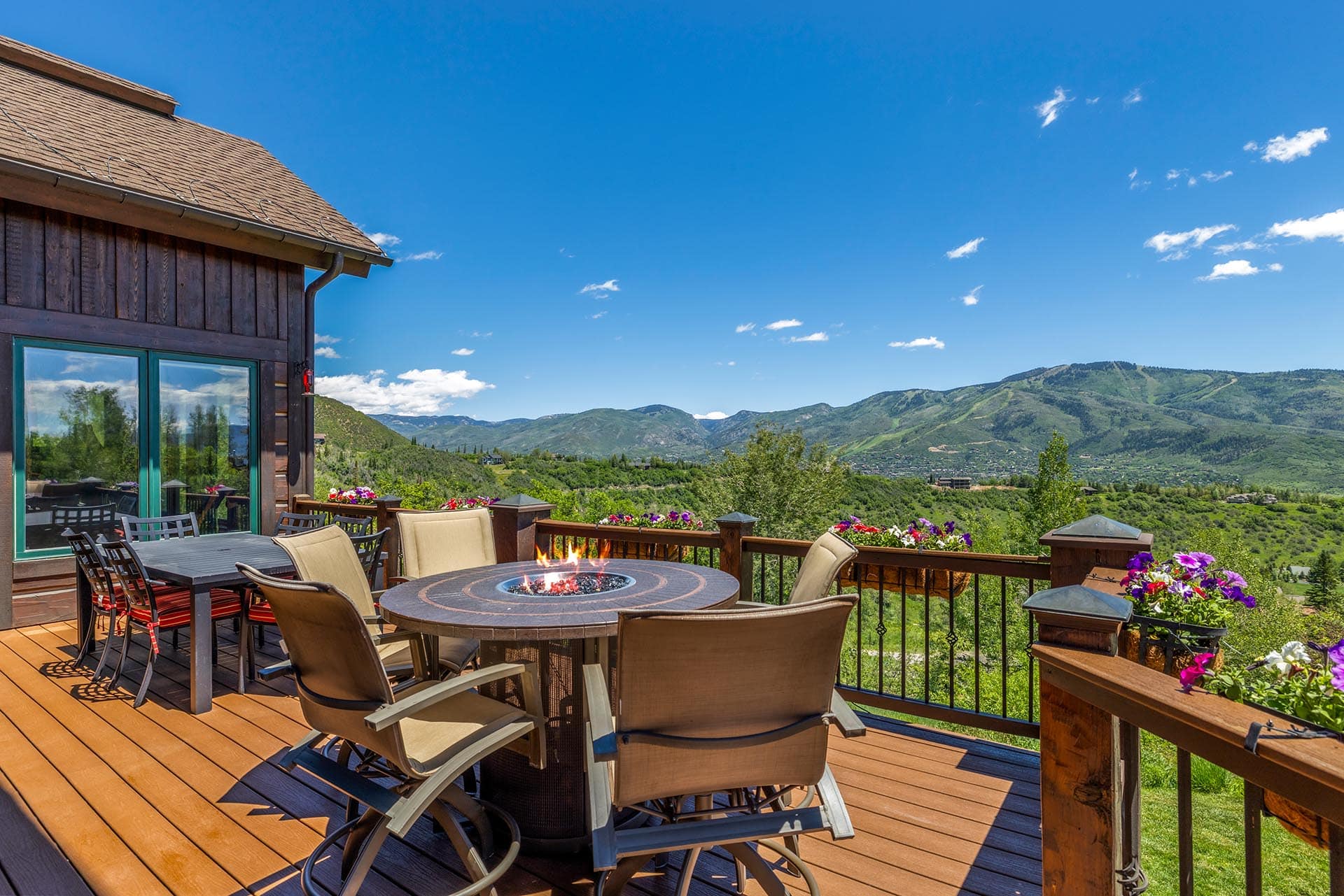 Outdoor wooden deck with dining table, chairs, and a central fire pit, overlooking green hills and distant mountains under a blue sky.