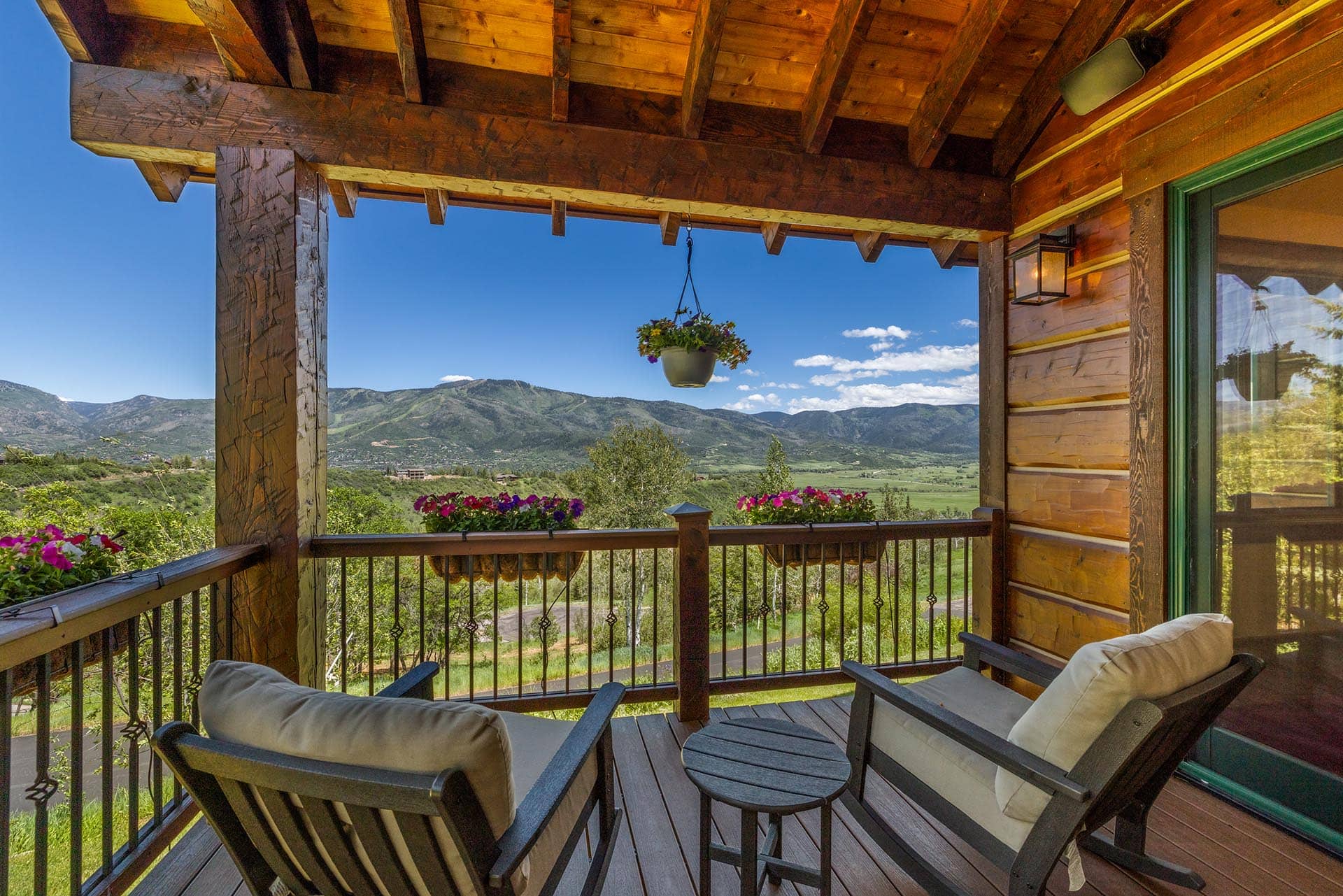 Cozy wooden balcony with cushioned chairs, small table, and a hanging flower pot overlooking a mountain valley.