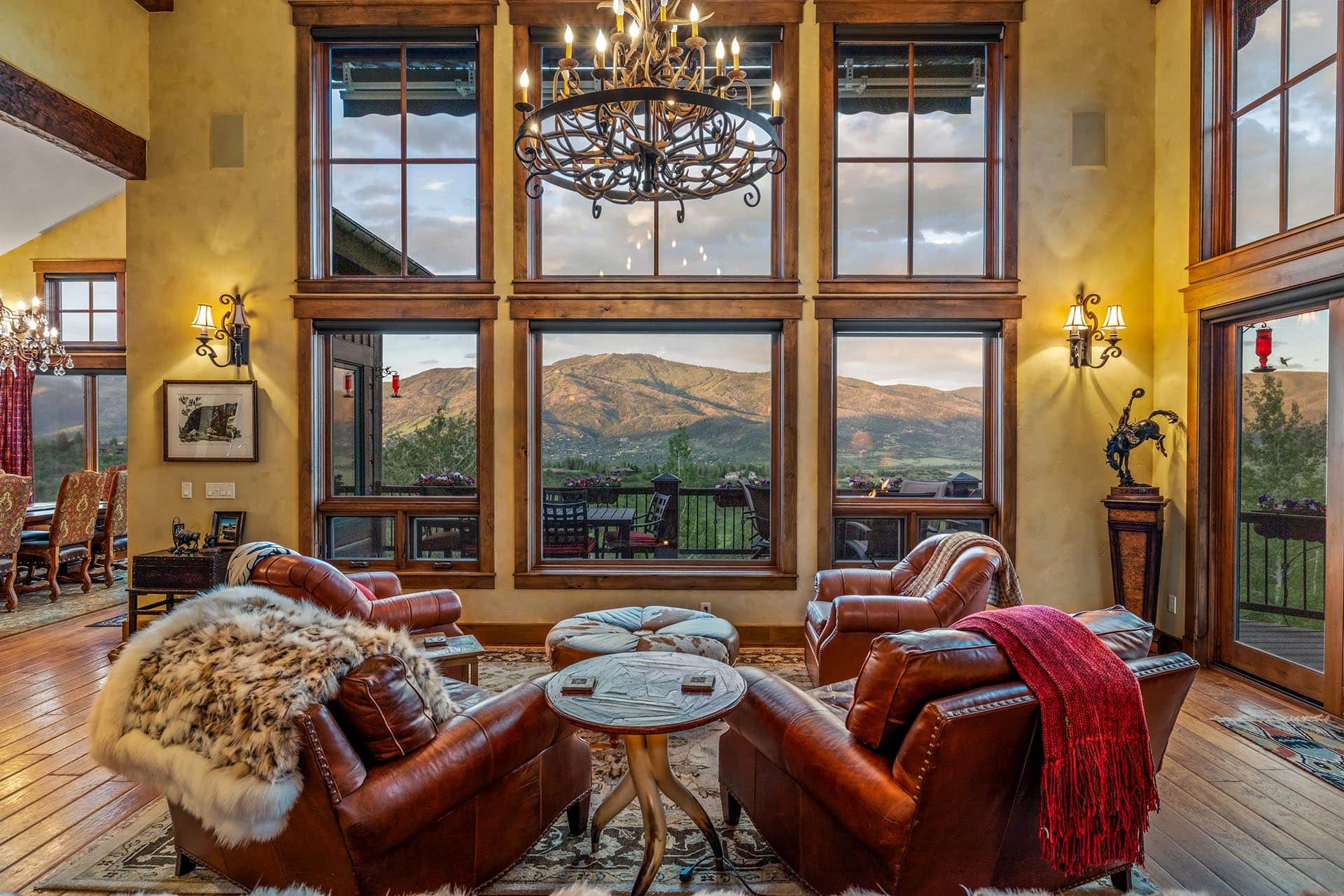 Cozy living room with brown leather chairs arranged around a round table, large wooden-framed windows showing a mountain landscape outside.