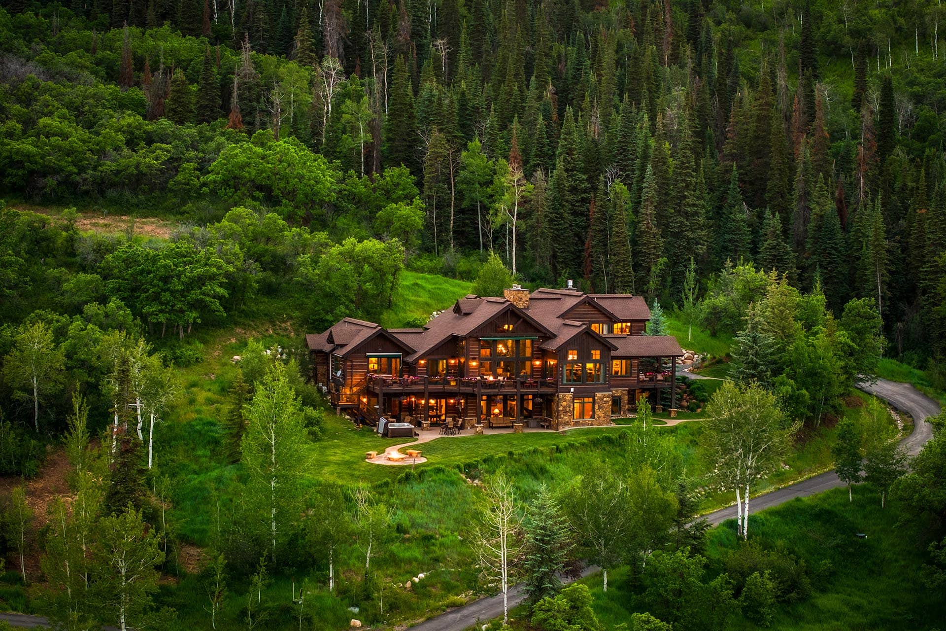 Large wooden cabin with warm lights nestled in a dense pine forest, with a curved driveway nearby.