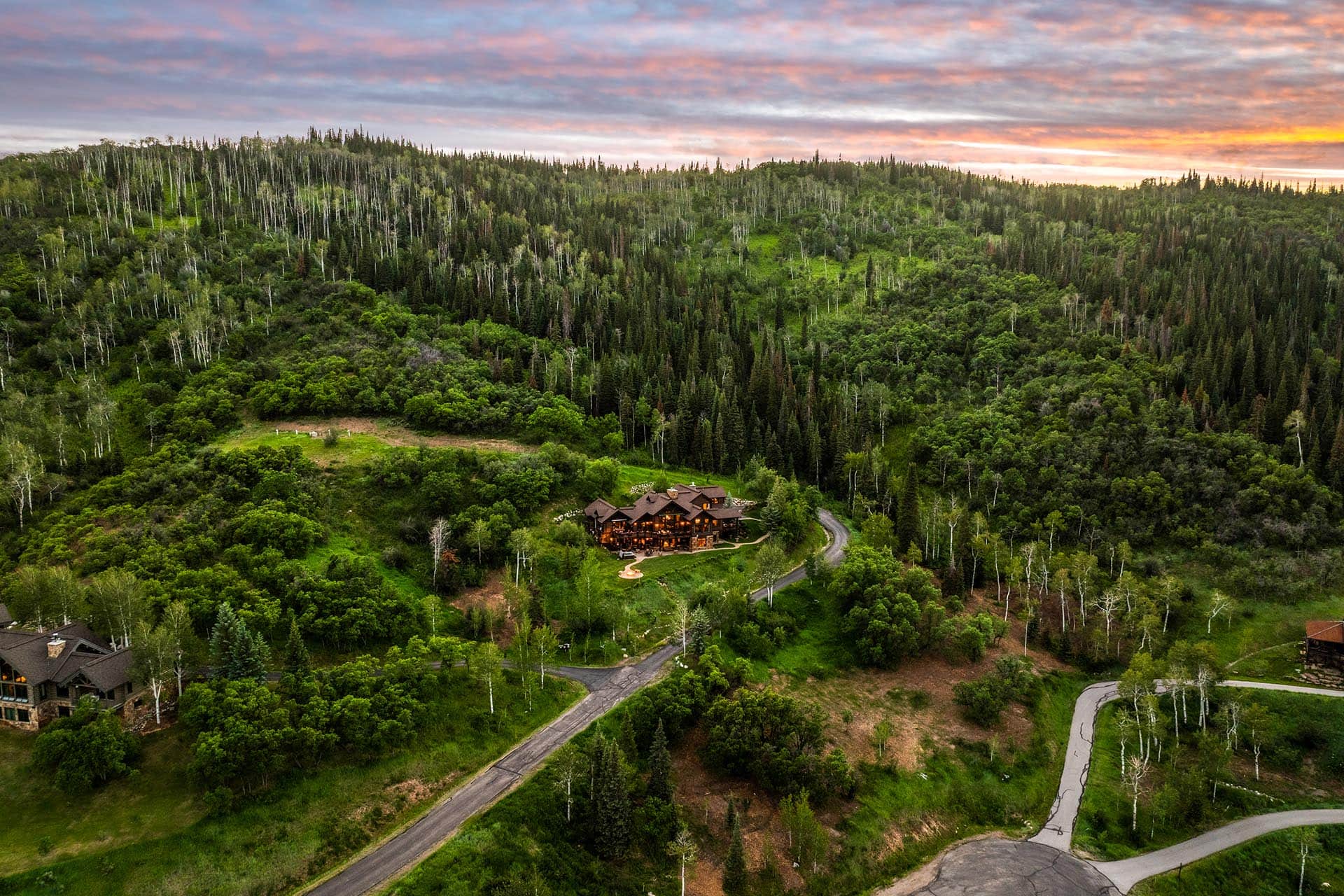 A large lodge nestled among dense green trees on a hillside with a winding road leading to it at sunset.