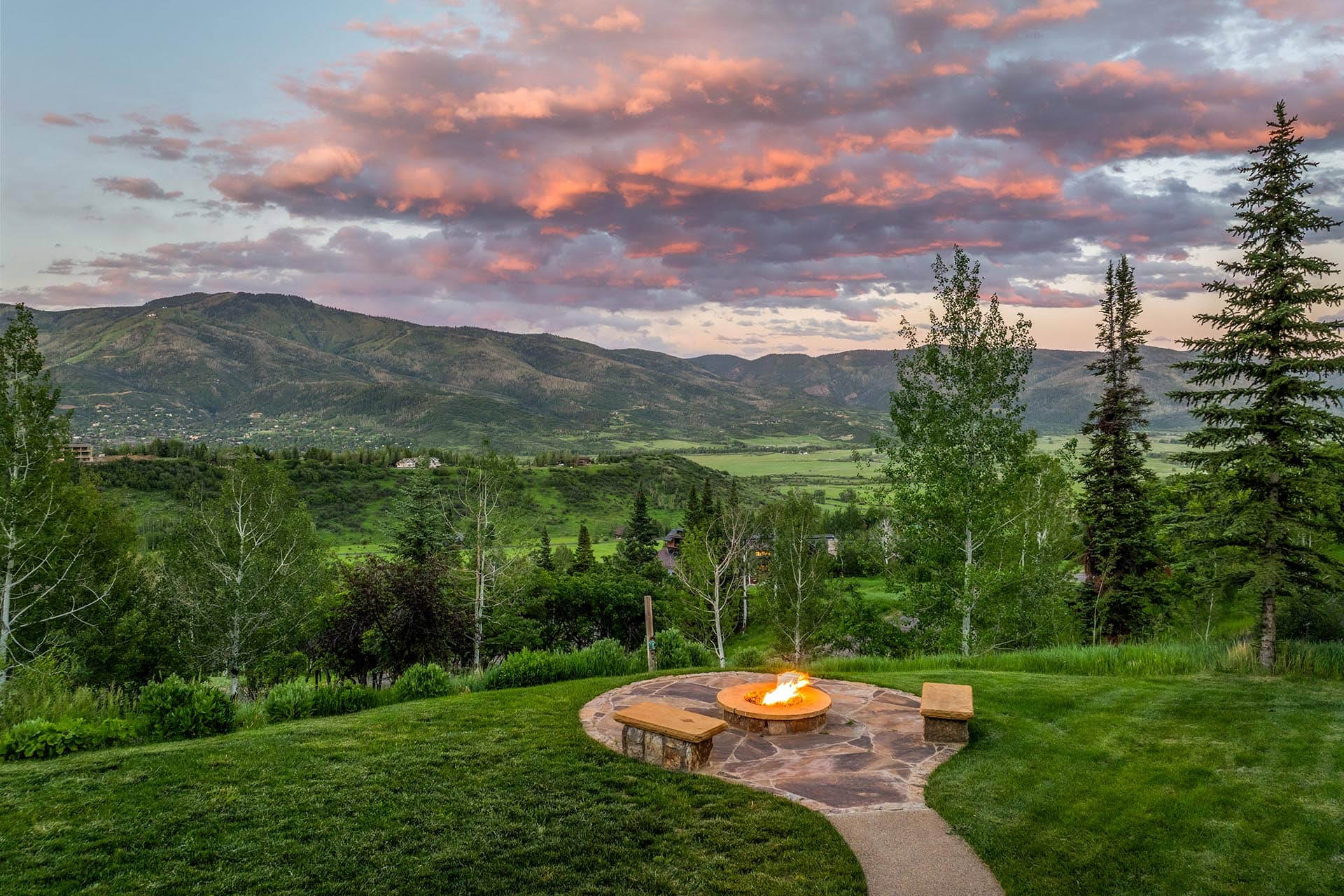 Sunset over a green valley with mountains and scattered trees; a lit stone fire pit sits in a landscaped yard in the foreground.