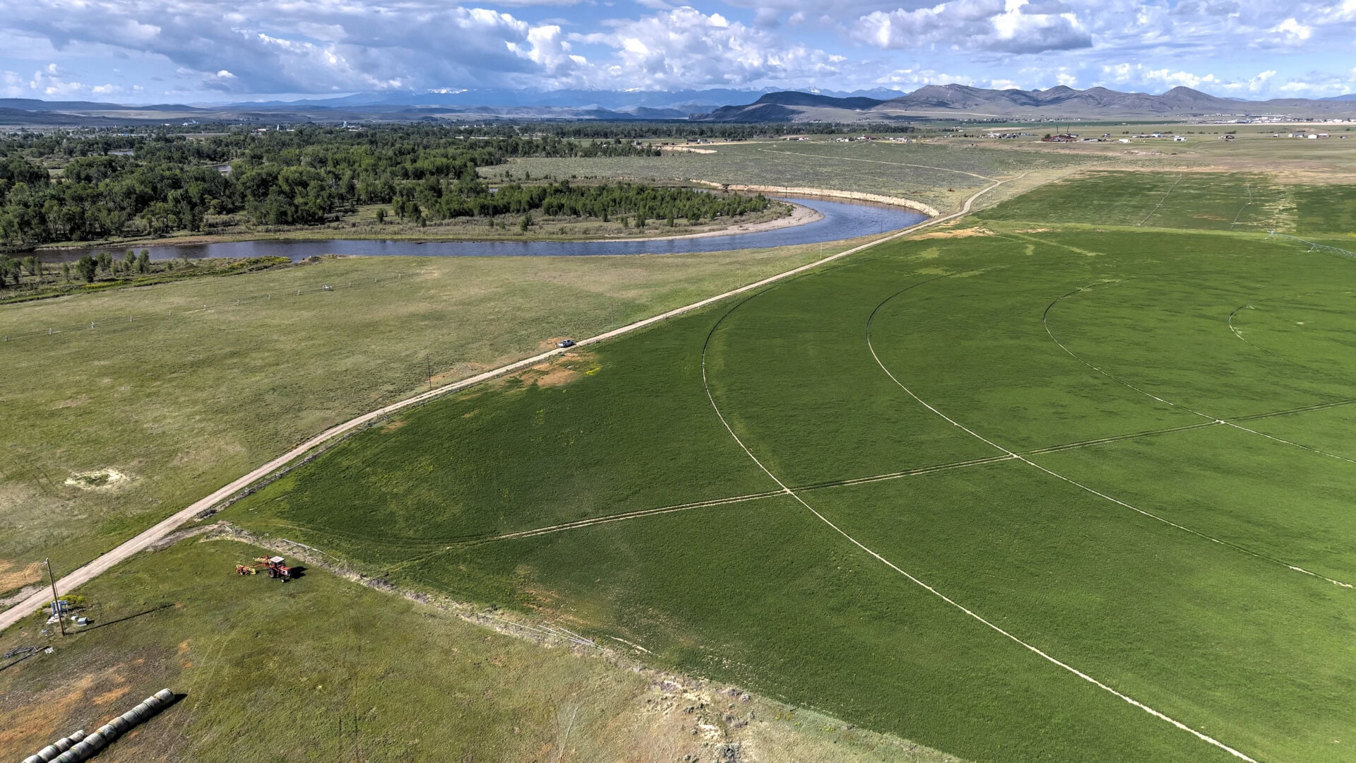 Aerial view of a winding river through green fields with a dirt road and distant mountains under a cloudy sky.