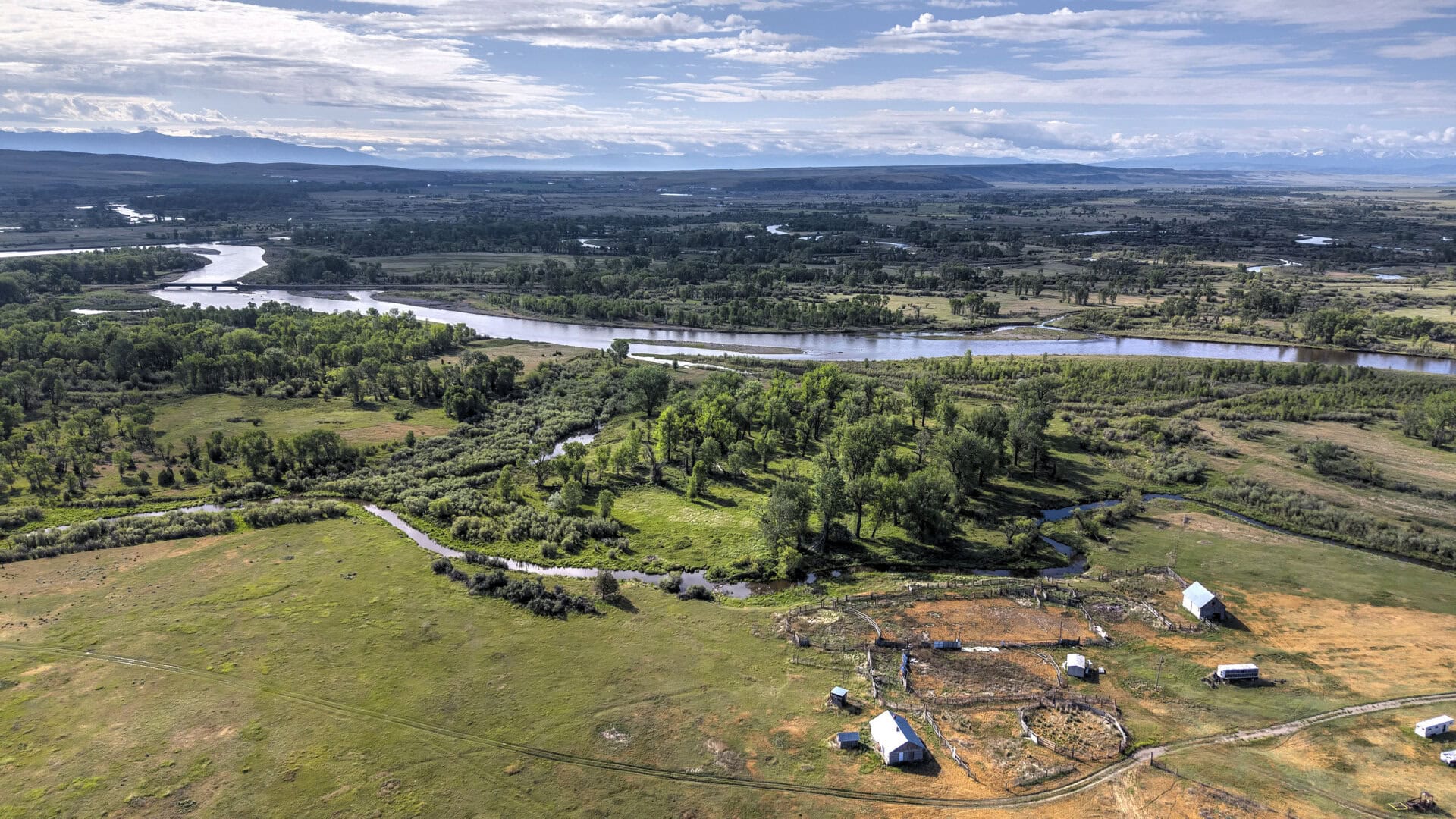 Aerial view of a winding river through a green valley with fields, trees, and distant mountains under a blue sky.
