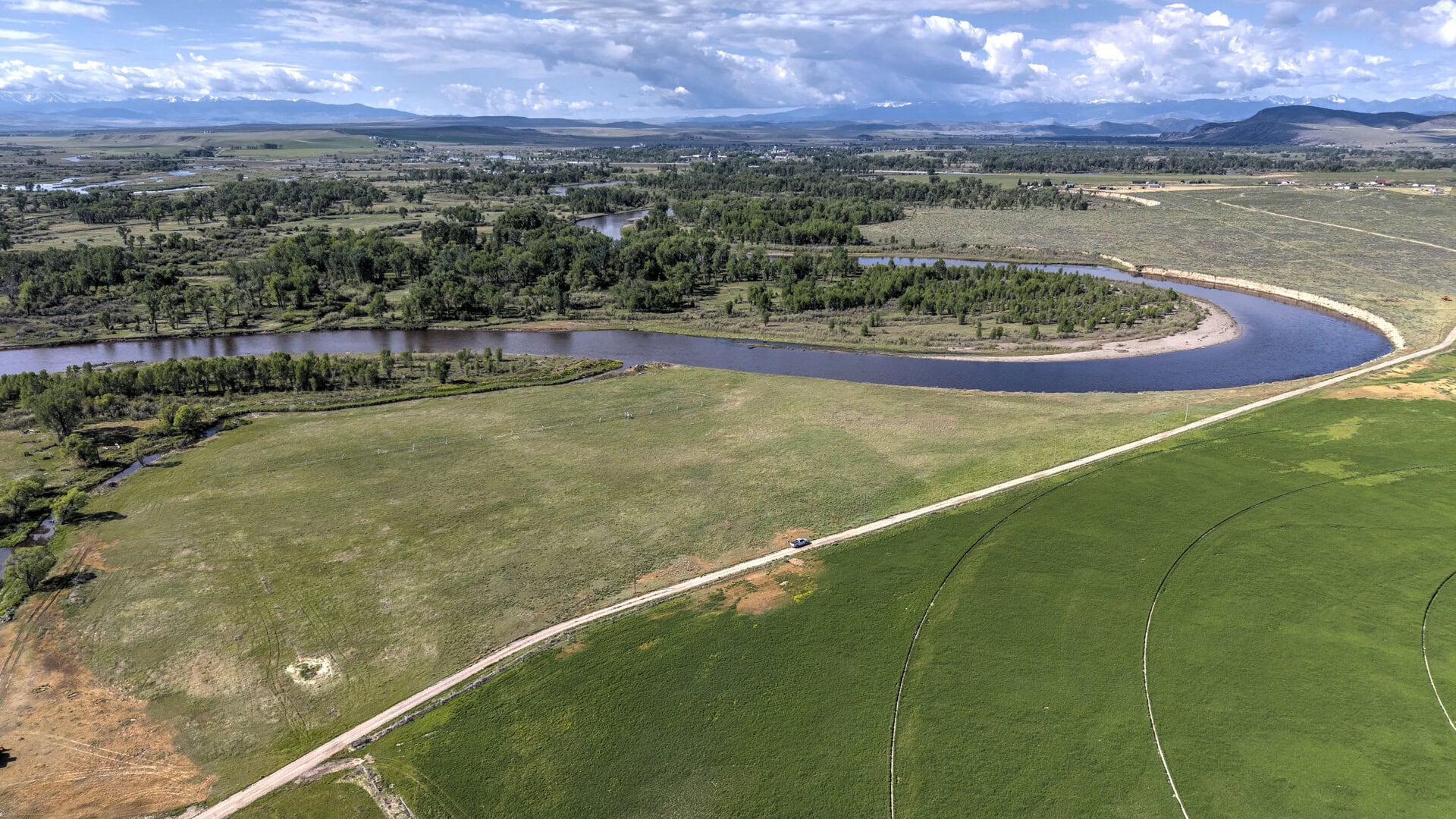 Aerial view of a winding river through green fields, with a dirt road, trees, and distant mountains under a partly cloudy sky.