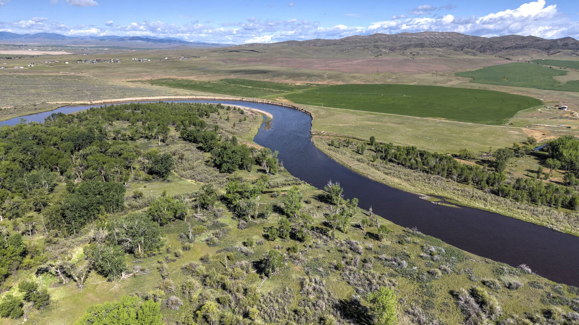Aerial view of a winding river curving through a grassy, tree-filled landscape with green fields and distant hills under a blue sky with clouds.