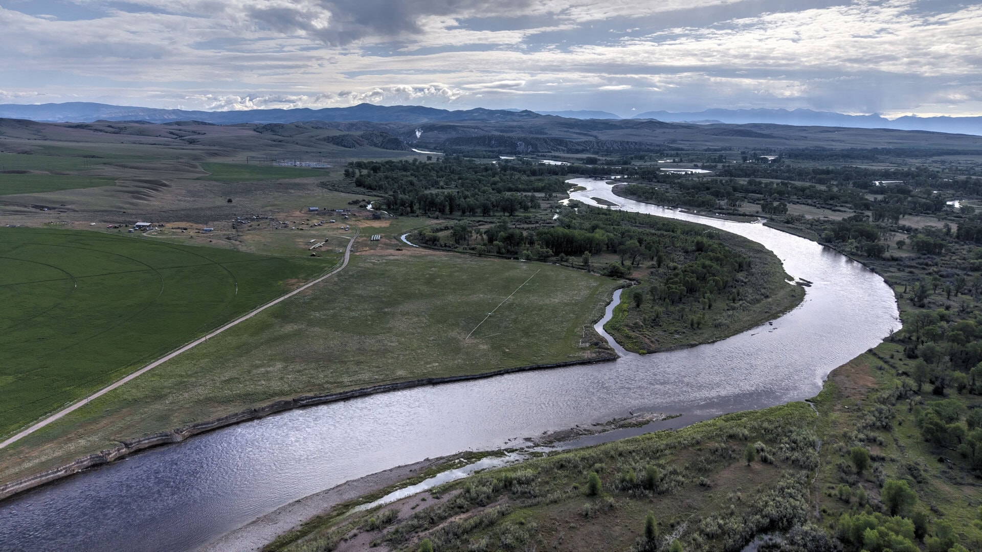 Aerial view of a winding river through green fields and scattered trees with distant mountains on the horizon, under a cloudy sky.