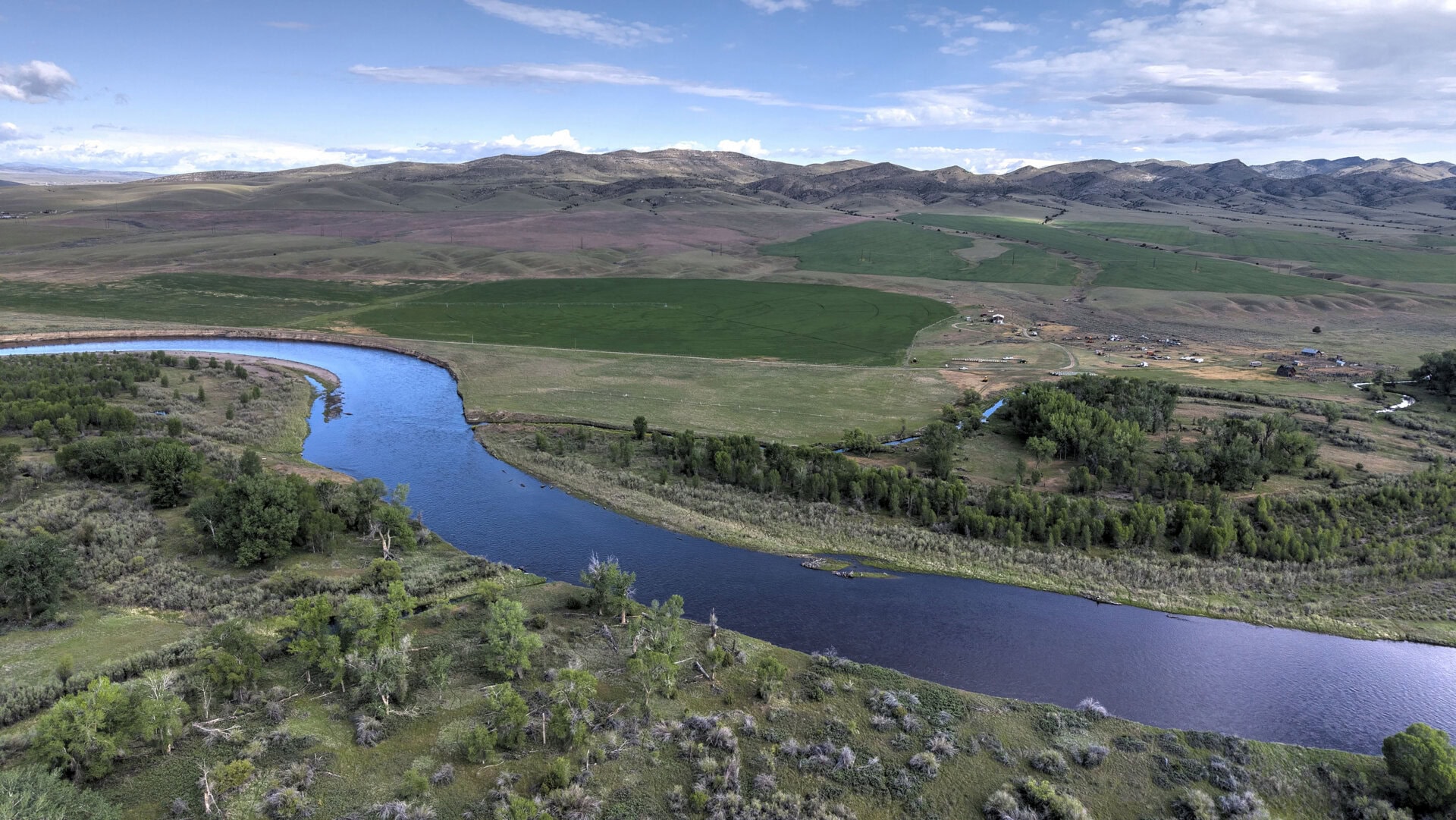 Aerial view of a winding river through green fields and rolling hills, with a small farm cluster on the right and trees in the foreground under a blue sky.