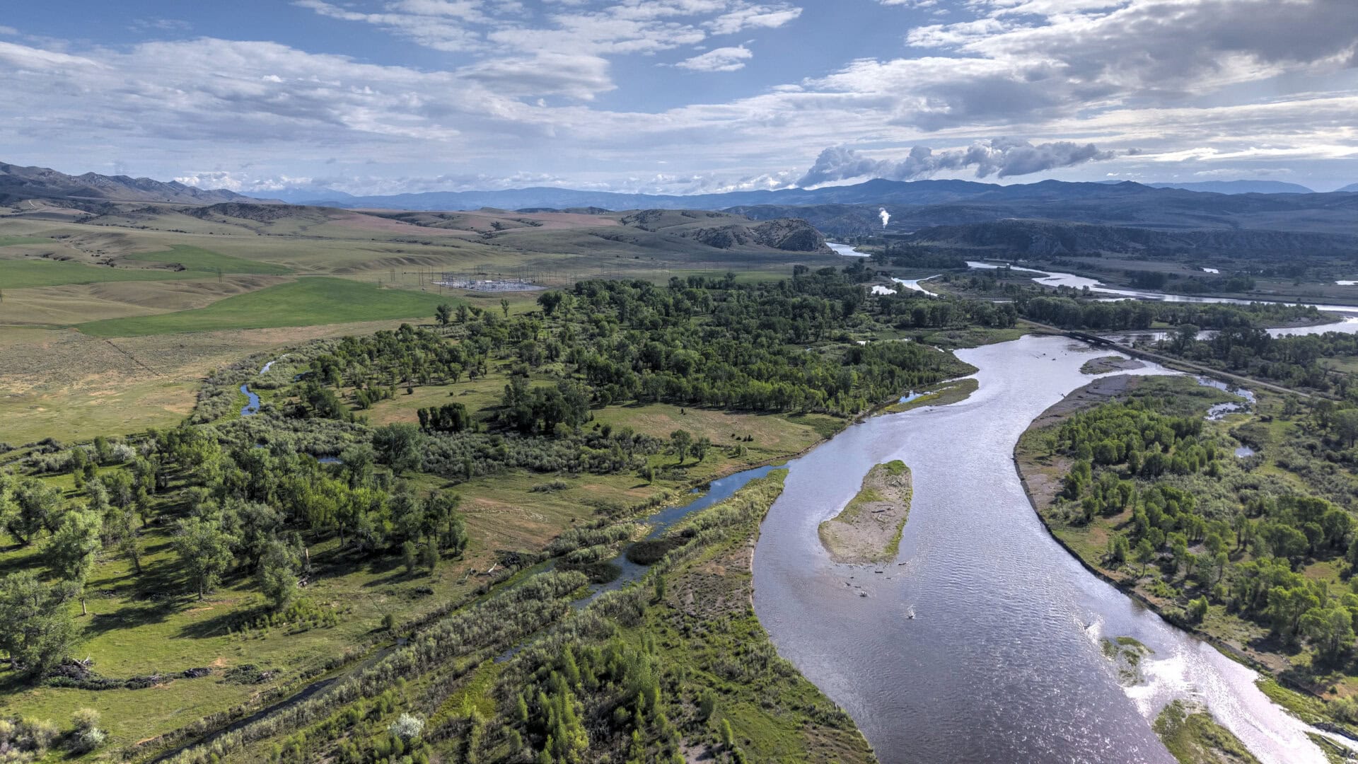 Aerial view of a winding river through green fields and wooded banks with rolling hills and distant mountains under a partly cloudy sky.