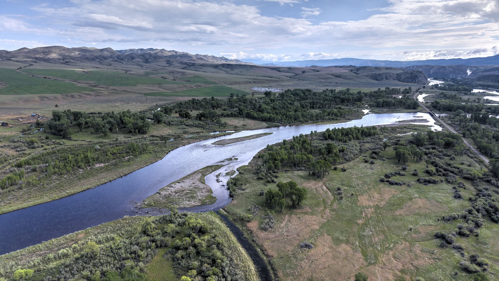 Aerial view of a winding river through green fields and a forested valley, with hills in the distance.