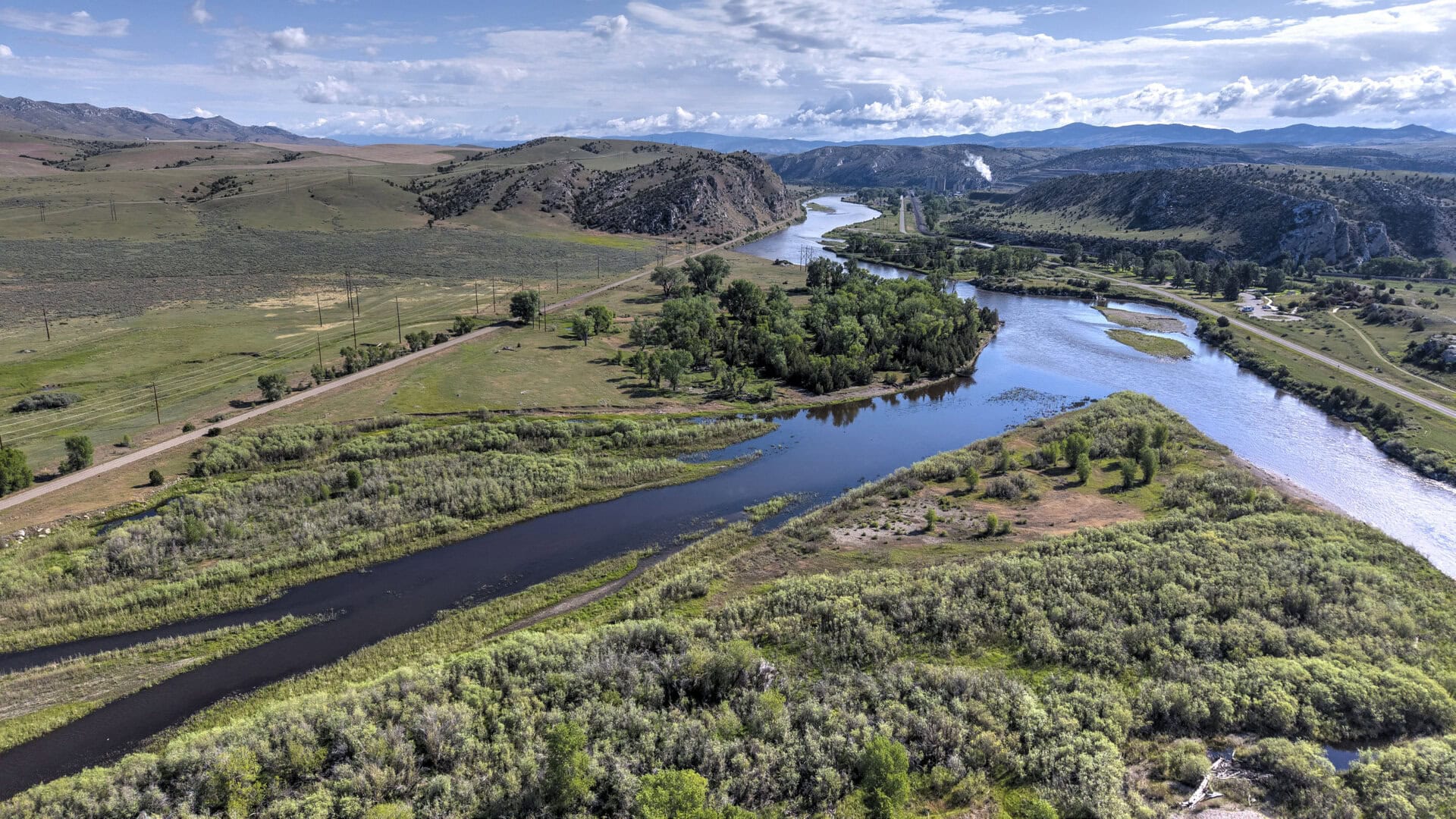 Aerial view of a winding river through green fields and rolling hills, with a road along the riverbank and clusters of trees on both sides.