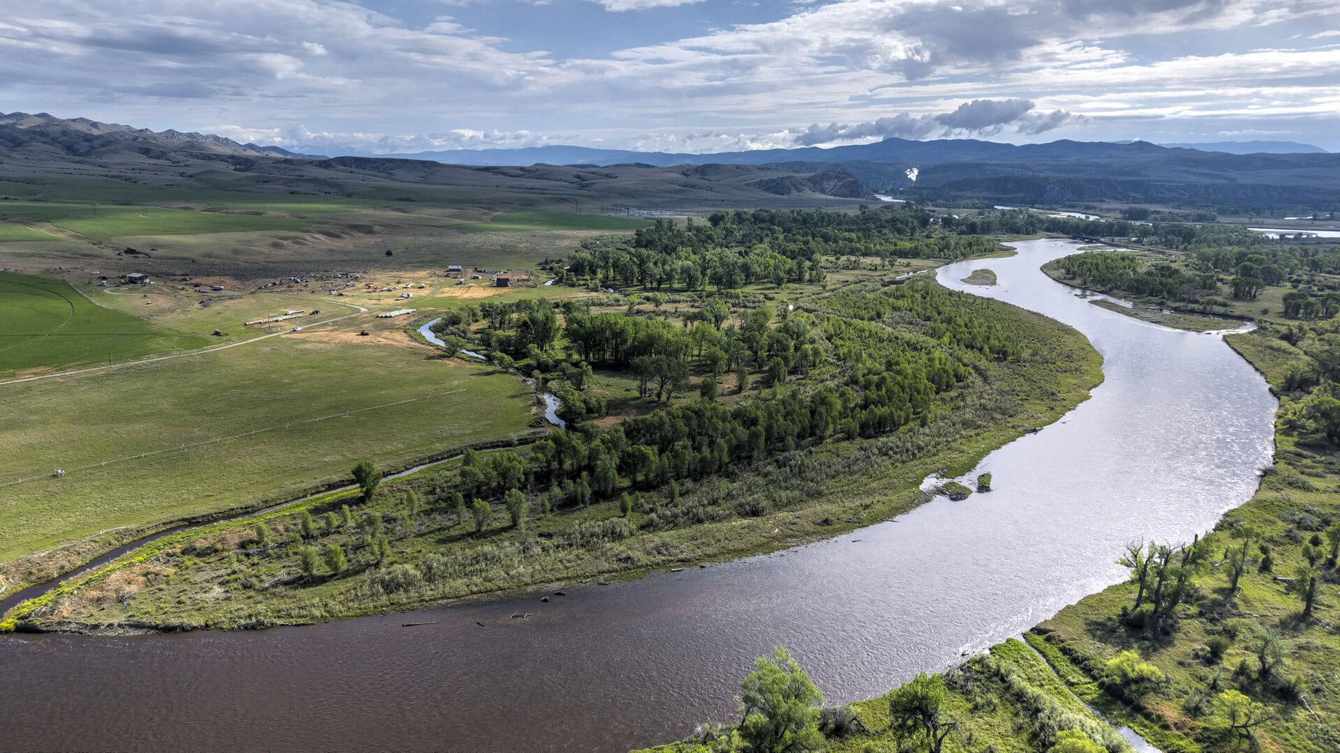 Aerial view of a winding river through green fields and trees, with hills and distant mountains under a cloudy sky.
