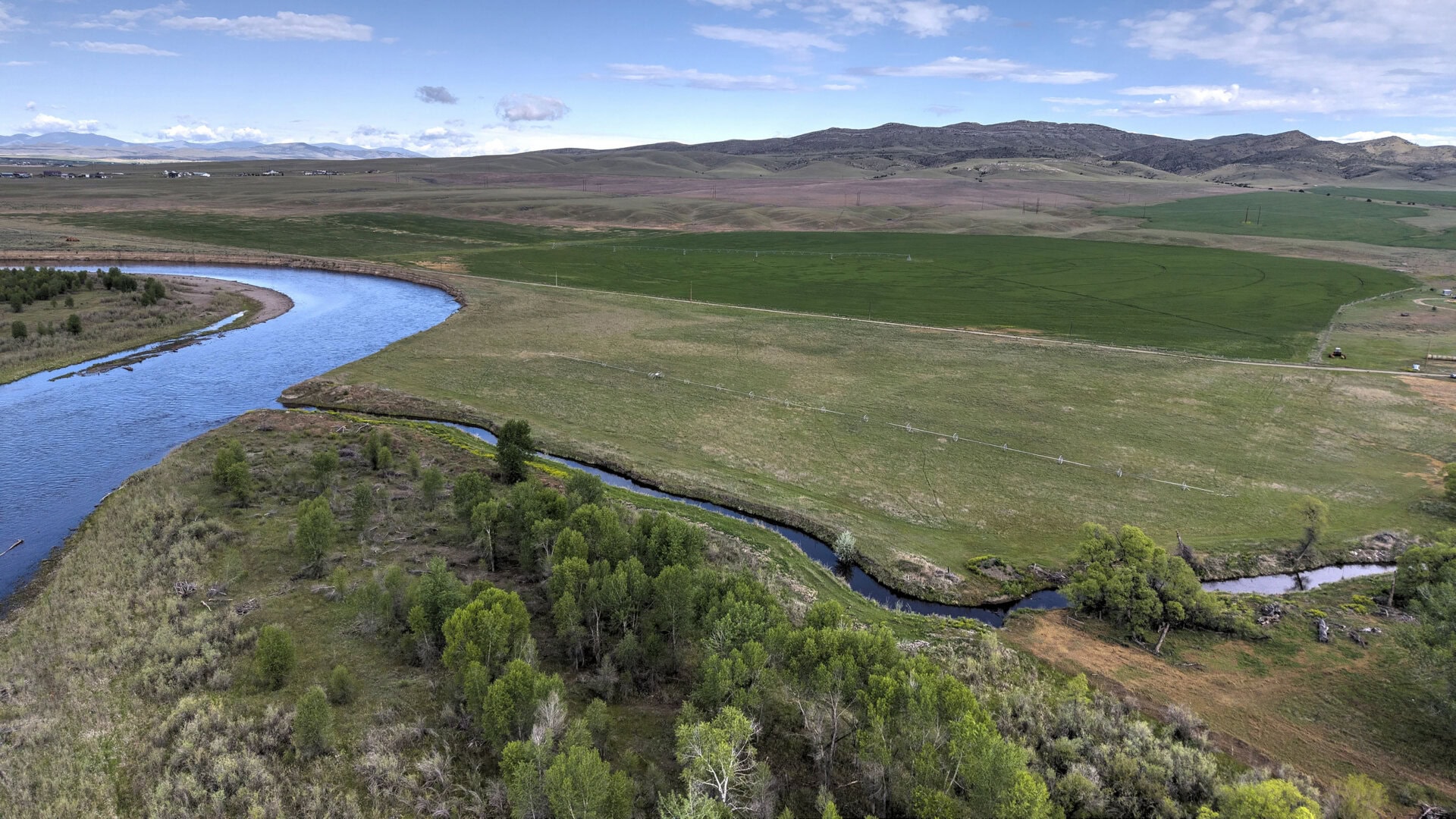 An aerial view of a winding river through green fields and distant rolling hills under a clear blue sky.