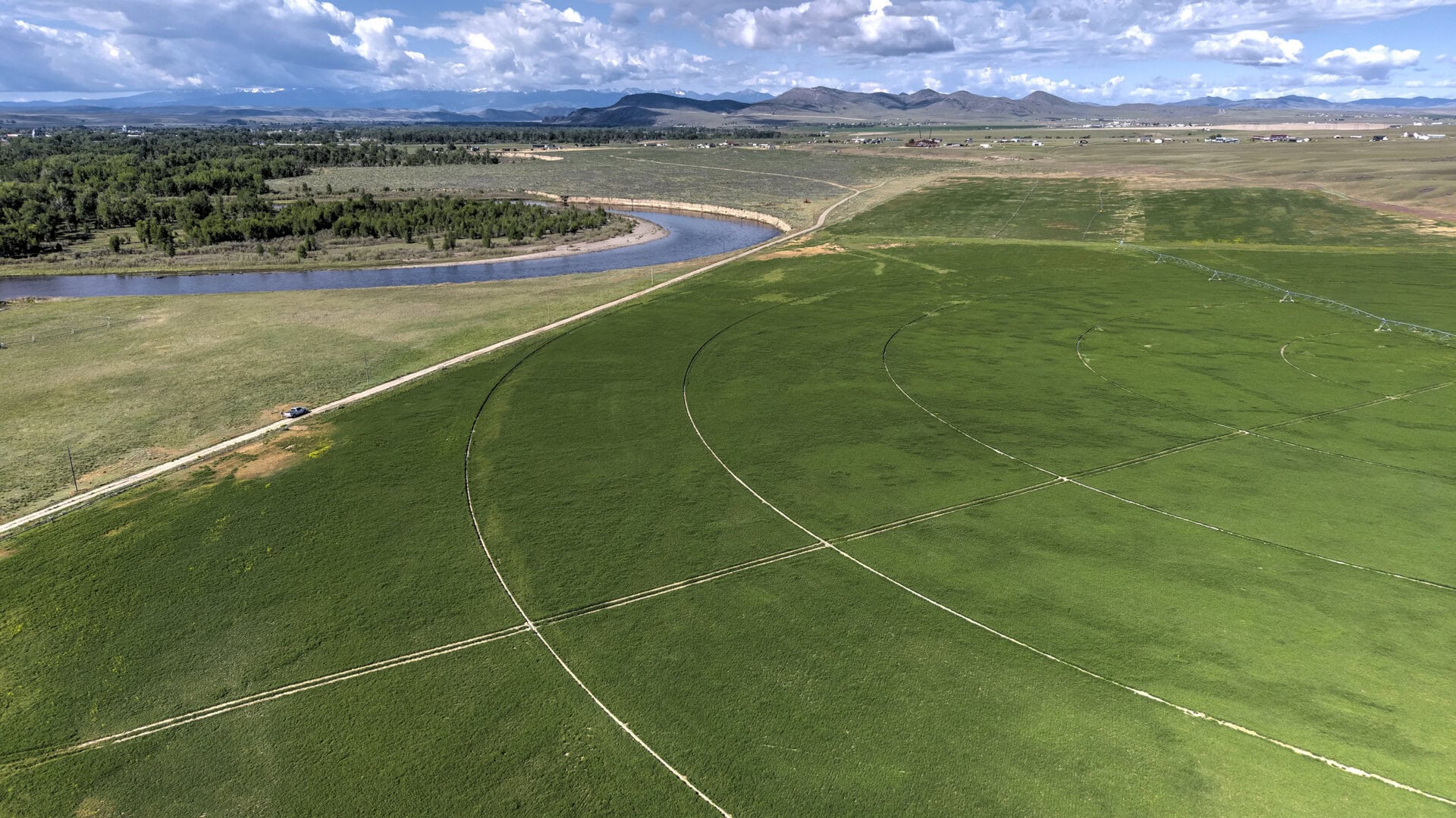 Aerial view of green fields with circular irrigation patterns, a winding road, and a river with trees along its banks, mountains in the distance.