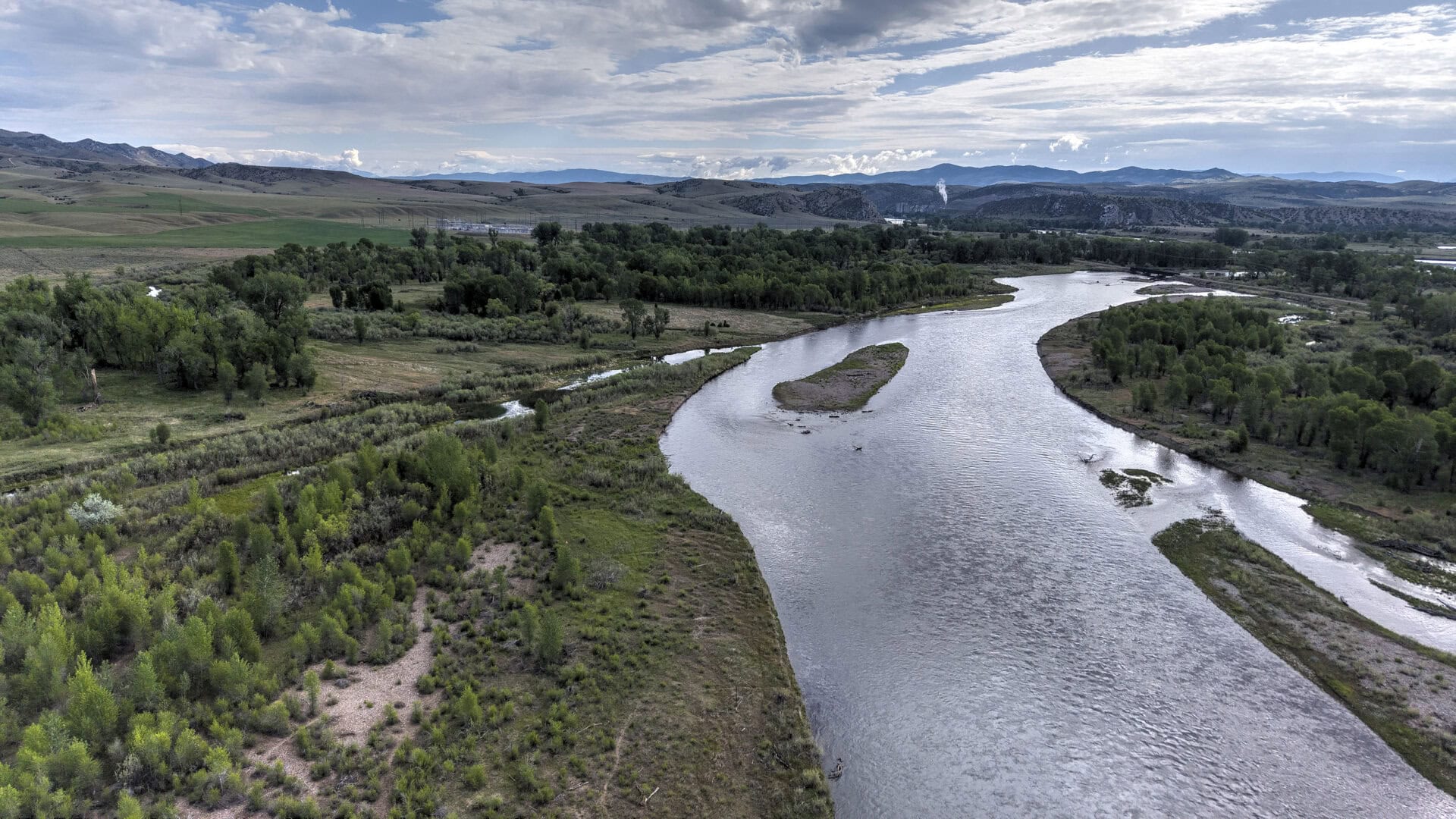 A winding river curves through a grassy valley with clusters of trees and distant mountains under a partly cloudy sky.