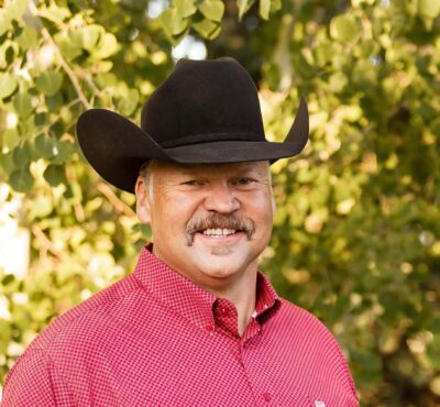 Smiling man wearing a black cowboy hat and red patterned shirt outdoors with green foliage in the background, crop from shoulders up