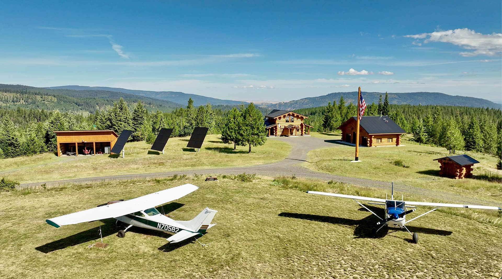 Small rural airfield with two small planes on a grassy field, wooden cabins, and a flagpole set against a forest and hills under a blue sky.
