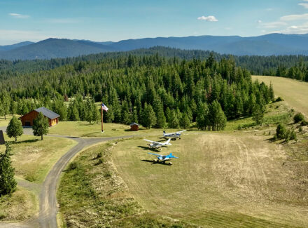 Aerial view of a rural airstrip with three small planes near a lodge, surrounded by pine forest and distant mountains.
