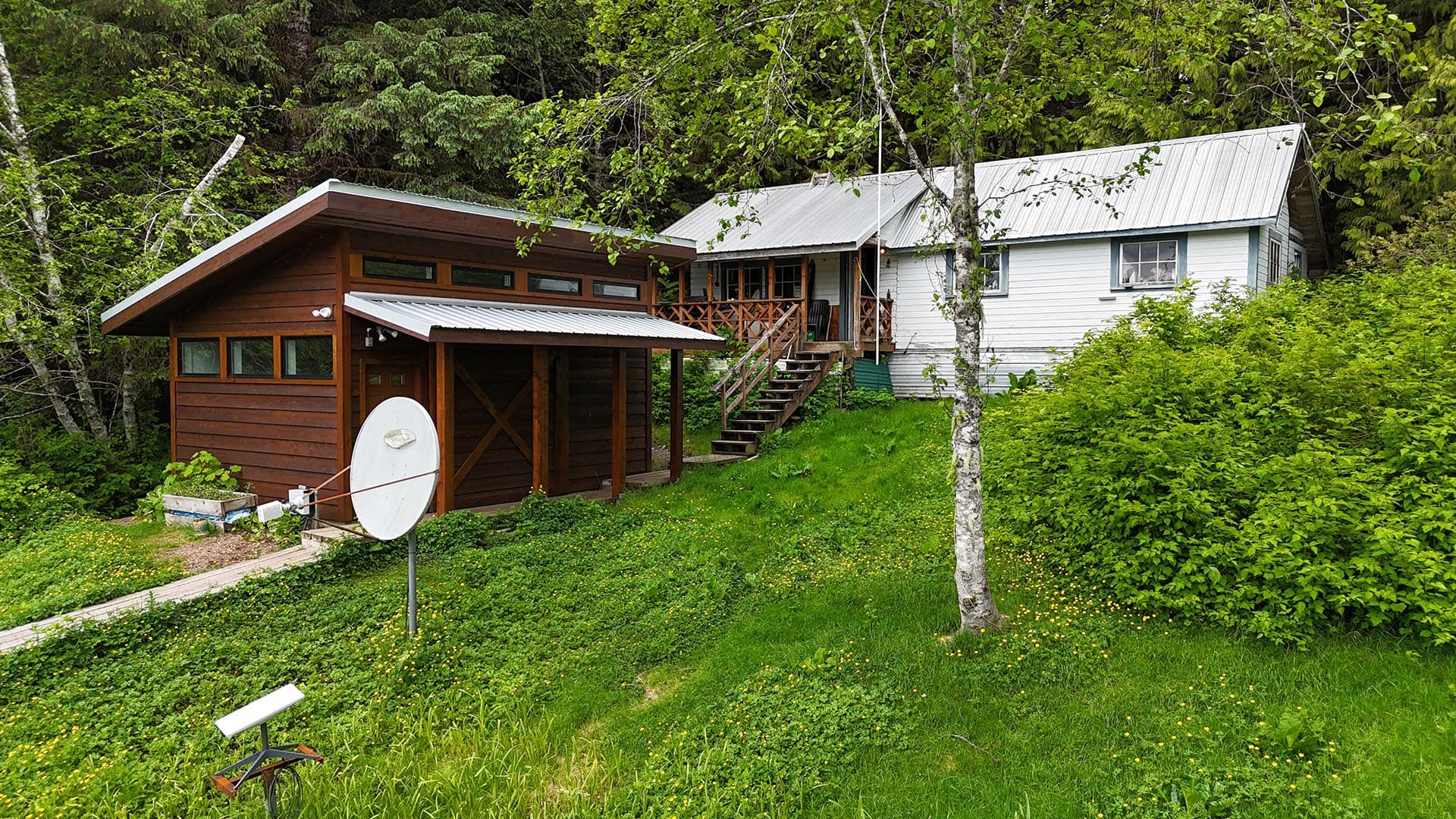 Brown wooden cabin attached to a white house, with a metal roof and wooden stairs, surrounded by green lawn and trees, satellite dish in foreground