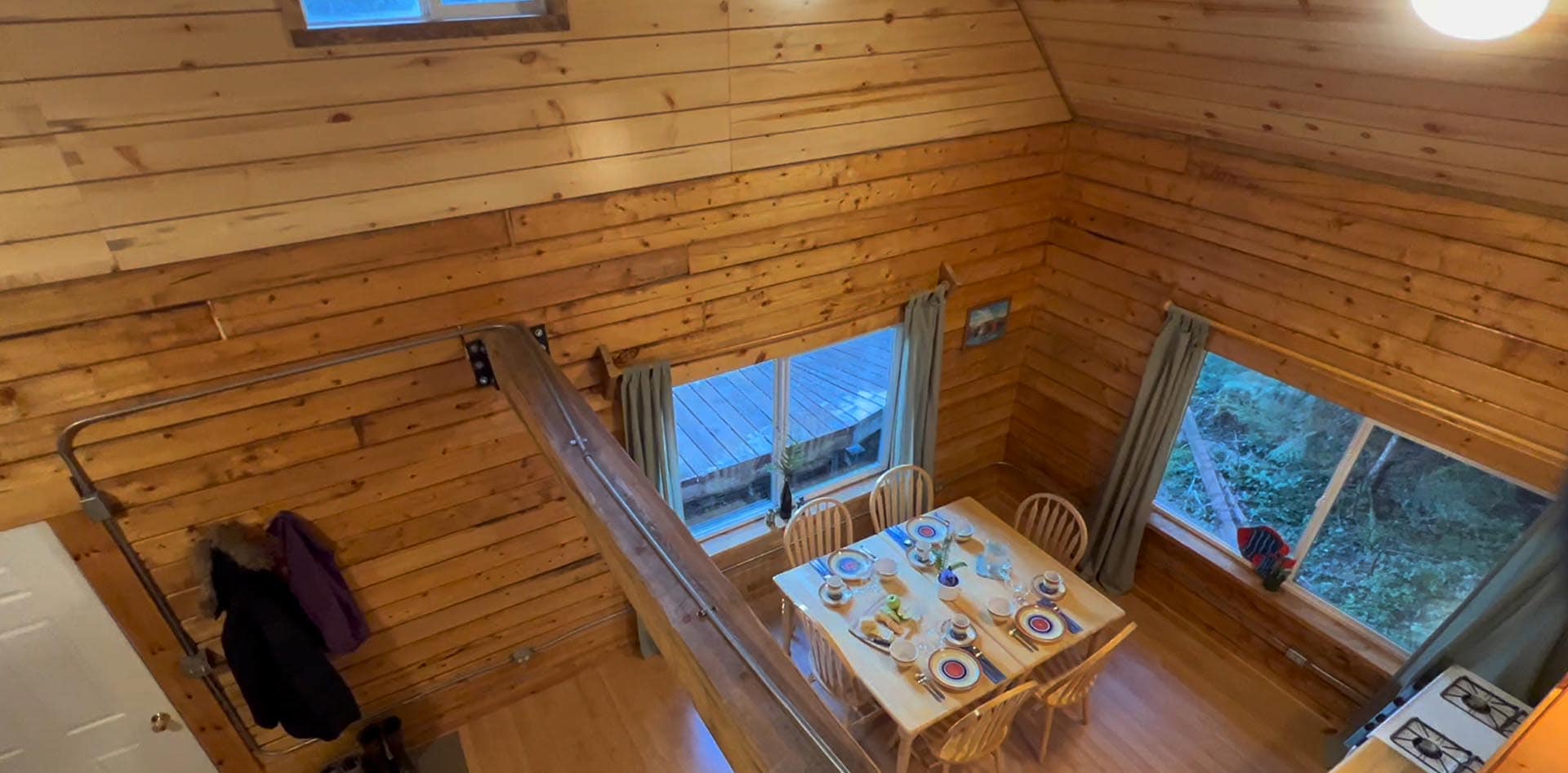 Overhead view of a pine cabin dining area with a long railing in the foreground and a table set for six near large windows