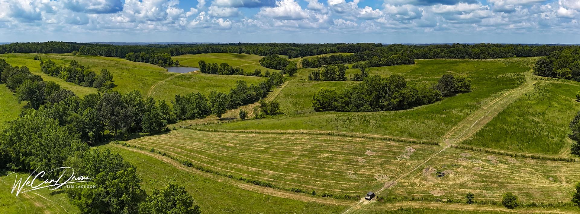 Aerial view of rolling green fields, trees, and a dirt road with a small vehicle, a pond in the distance under a cloudy sky.