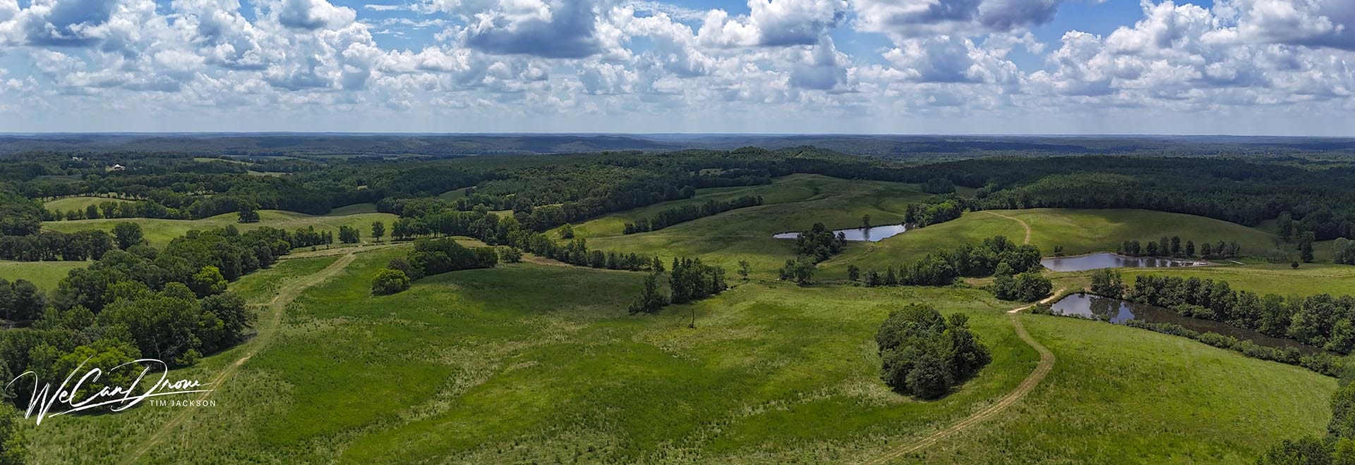 Aerial panoramic view of green rolling fields, ponds, and a winding dirt road under a blue sky with fluffy clouds.