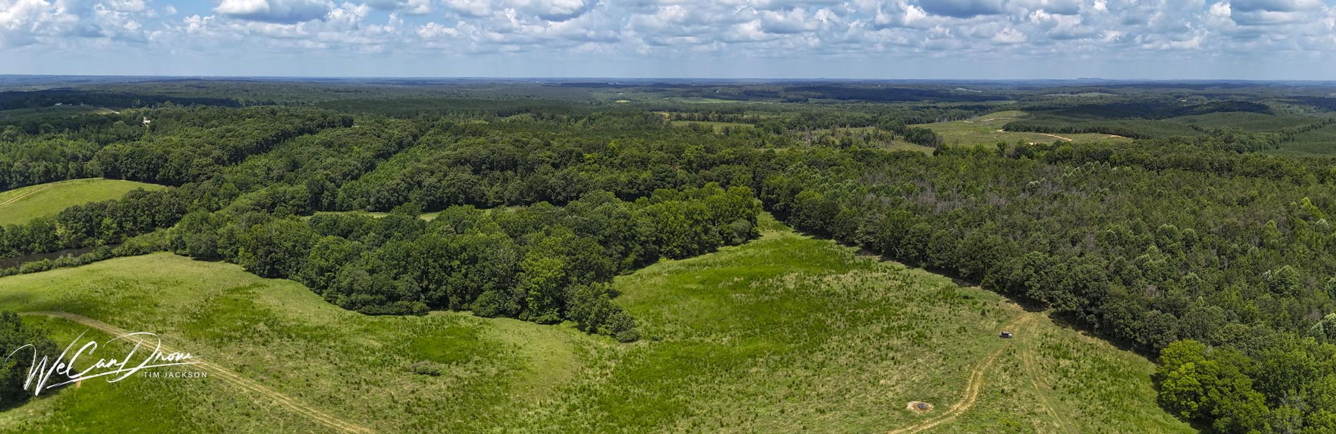 A panoramic aerial view of a lush green countryside with forests, open meadows, and distant hills; watermark in the lower-left corner.