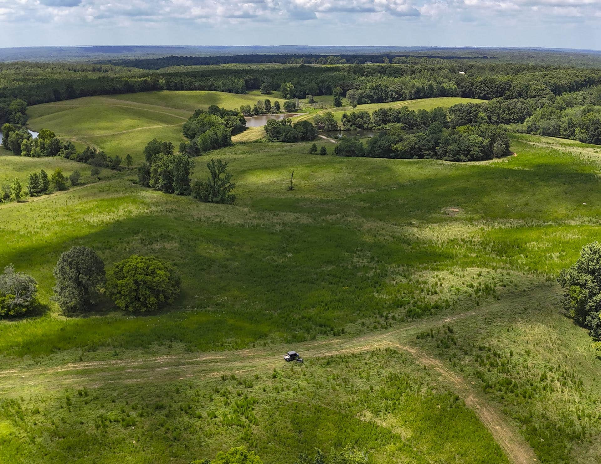 Aerial view of rolling green fields and forests with a dirt road and a small truck in the foreground.