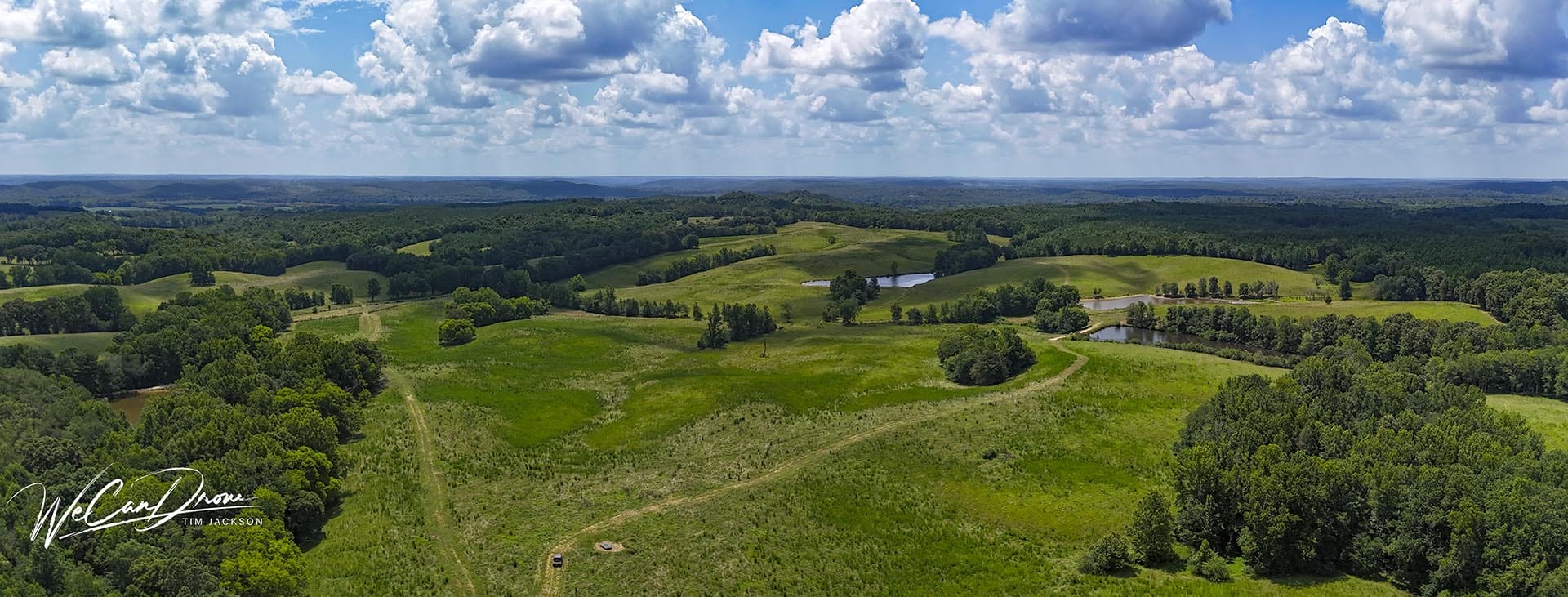 Aerial view of rolling green fields, trees, and small ponds under a bright blue sky with fluffy clouds.3d94fän11ata corners.