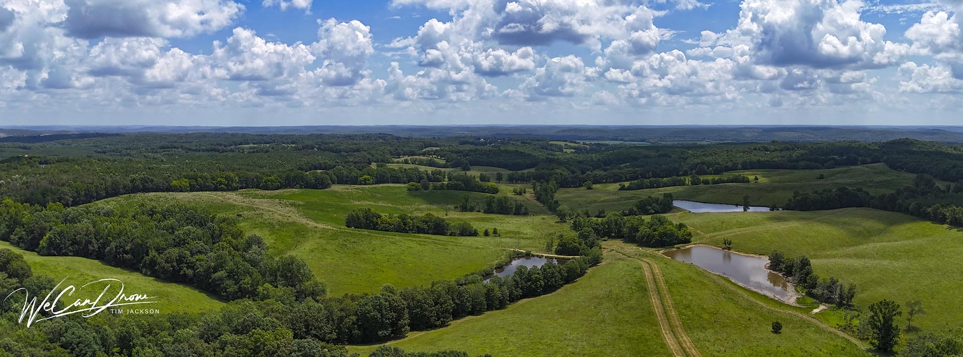 Aerial view of rolling green fields, trees, and small ponds under a blue sky with fluffy clouds; a rural landscape panorama. Signature watermark bottom-left reads 'We Can Draw'.
