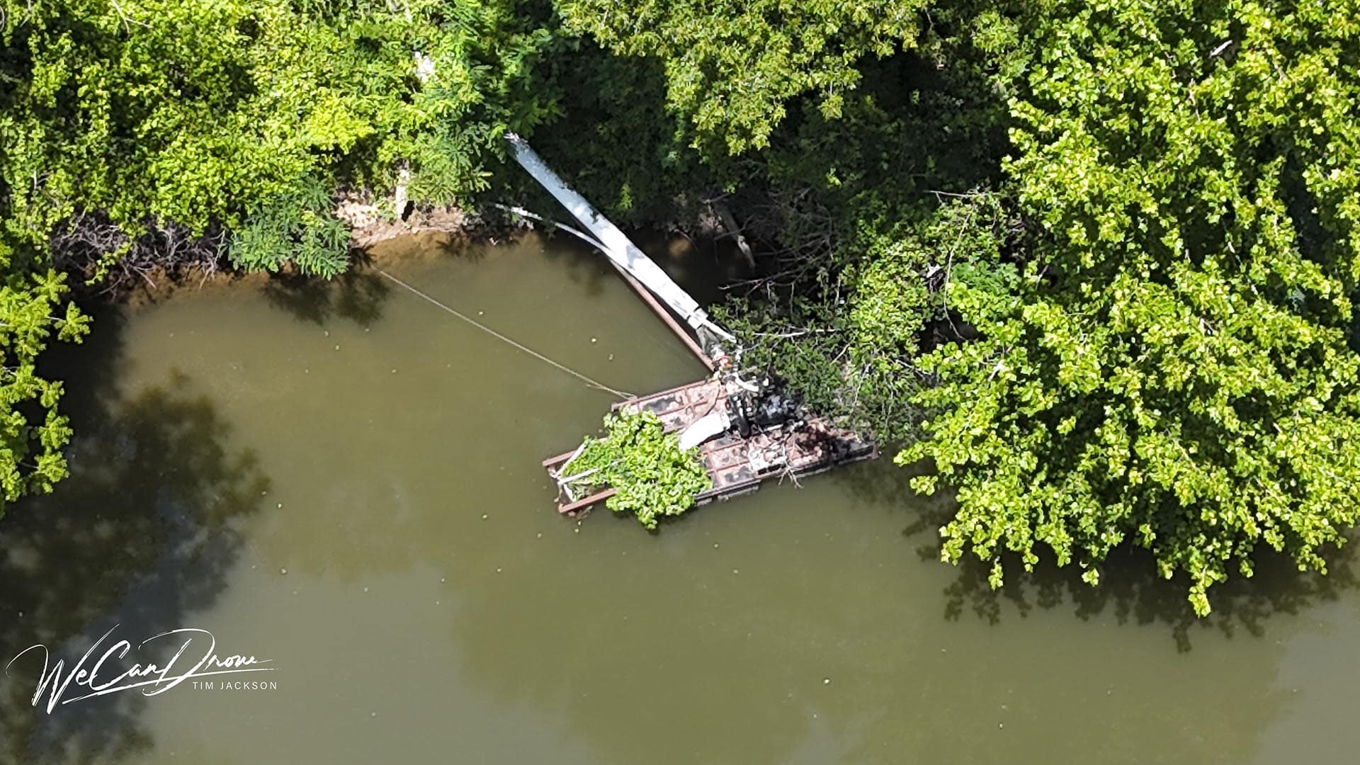 Aerial view of a partially submerged, rusted boat tangled in a riverbank beneath dense green foliage near the water's edge.