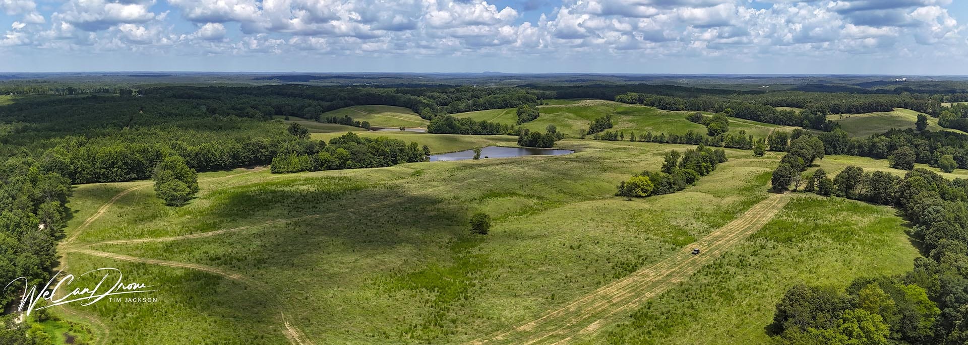Aerial view of rolling green fields, clusters of trees, and a small pond under a blue, cloud-filled sky.