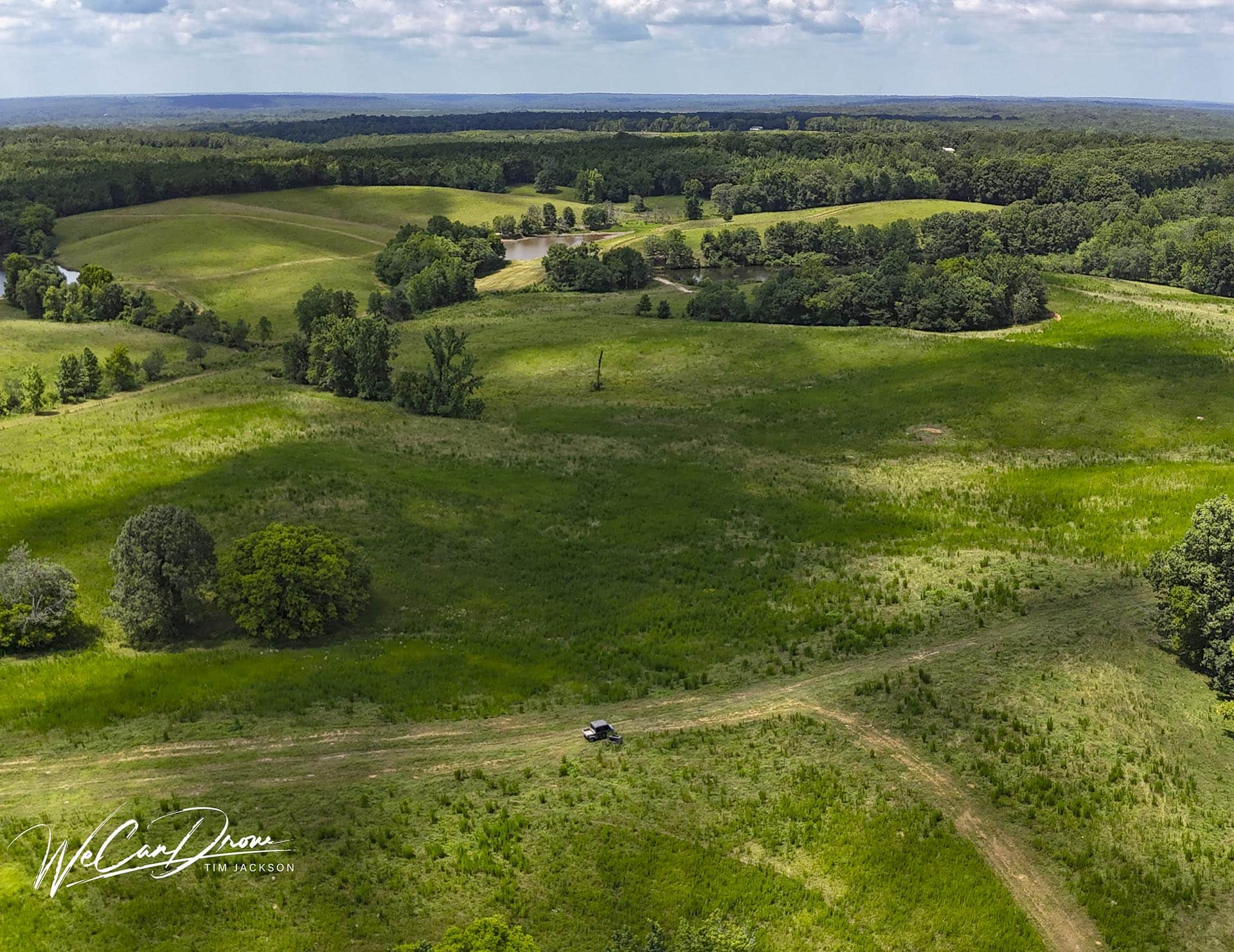 Aerial view of rolling green hills, clusters of trees, and a small pond, with a lone pickup truck on a dirt road in the foreground.