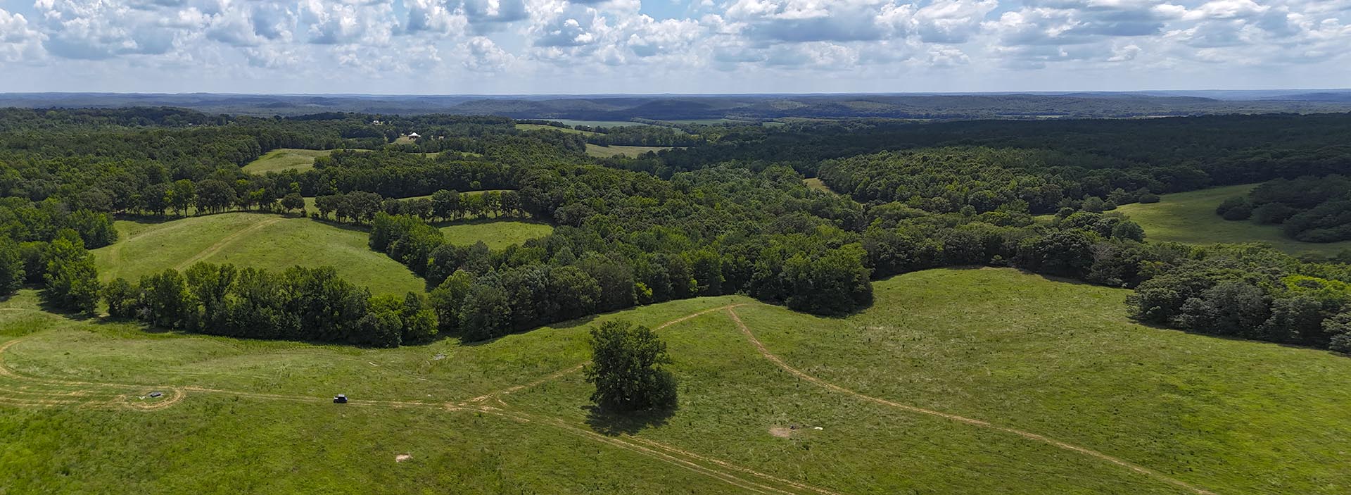 Aerial view of rolling green fields bordered by dense woodlands, with dirt paths and a lone vehicle.