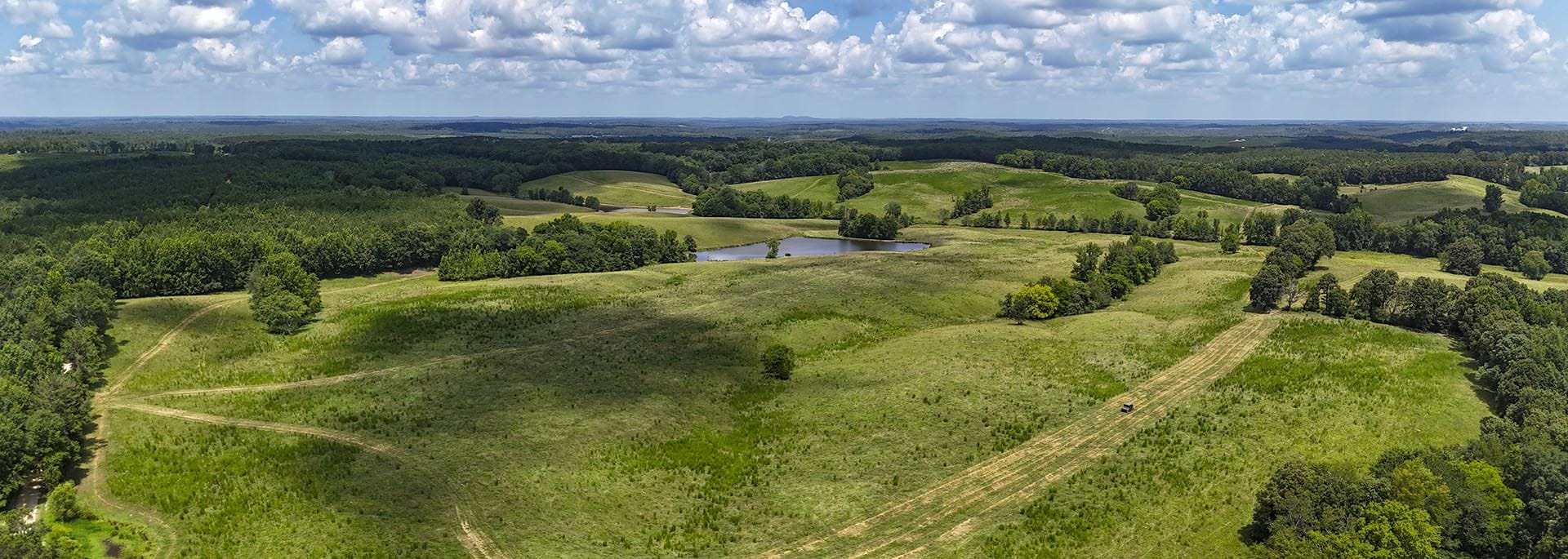 Panoramic aerial view of rolling green meadows, a small pond, trees, and dirt roads under a cloudy sky.