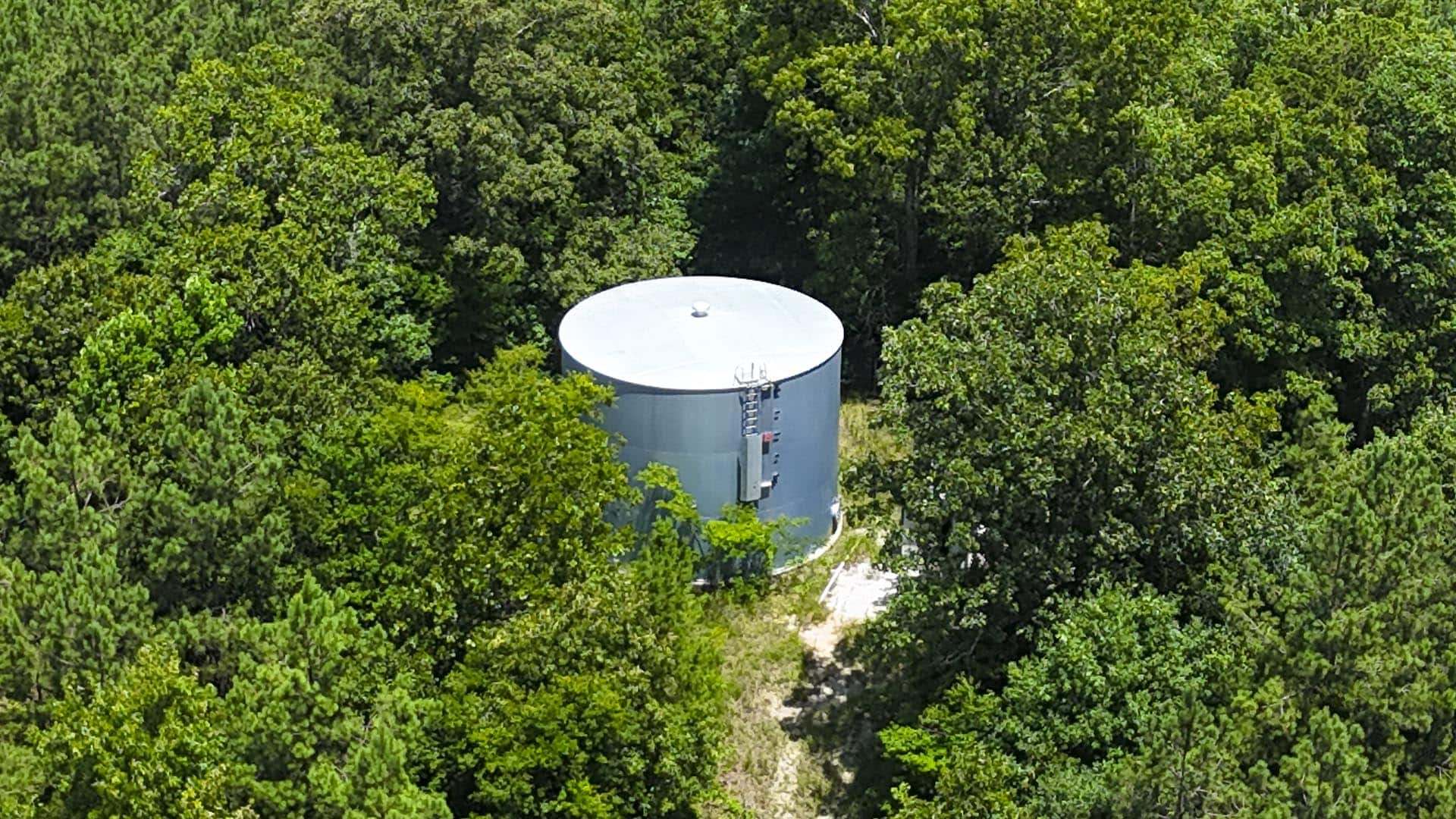 A cylindrical gray water storage tank sits in a clearing among dense green trees.