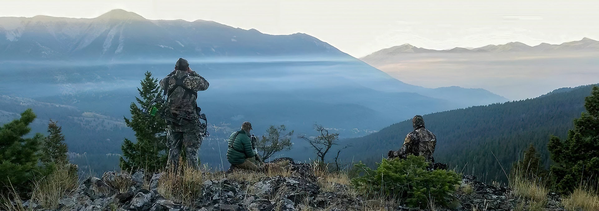 Three hunters in camouflage stand and sit on a rocky ridge, gazing over a foggy mountain valley at dawn.