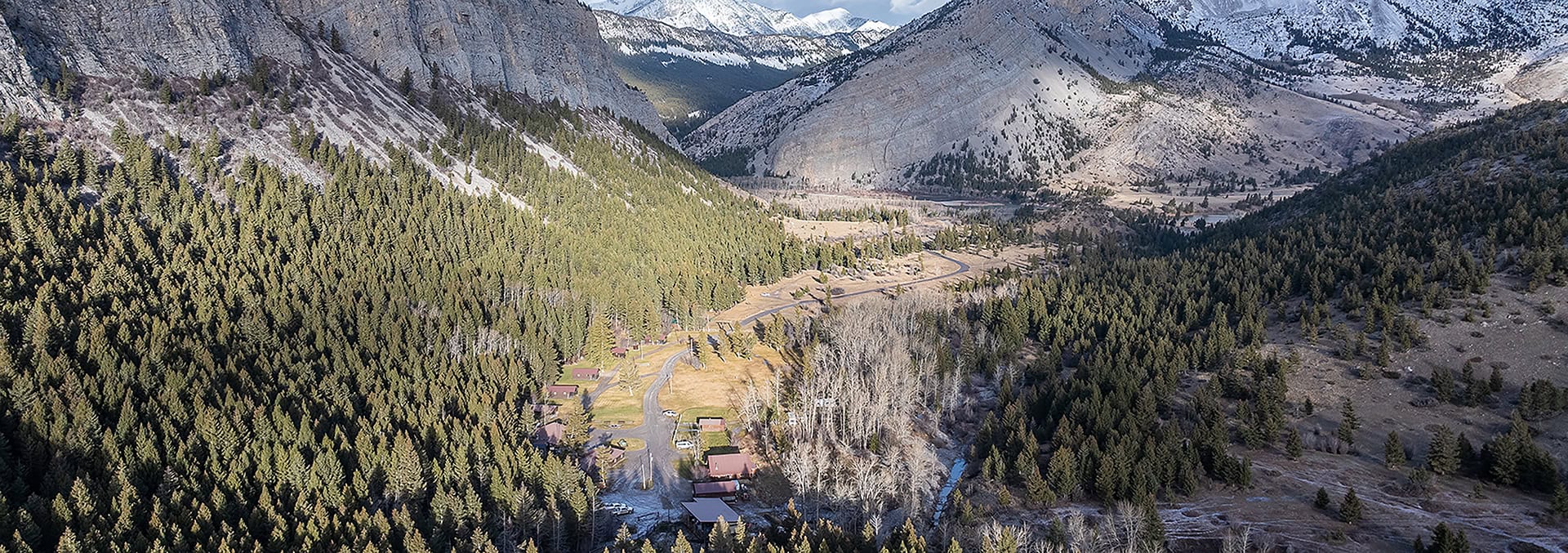 Aerial view of a rural village in a forested mountain valley with a winding road and snow-capped peaks in the distance.