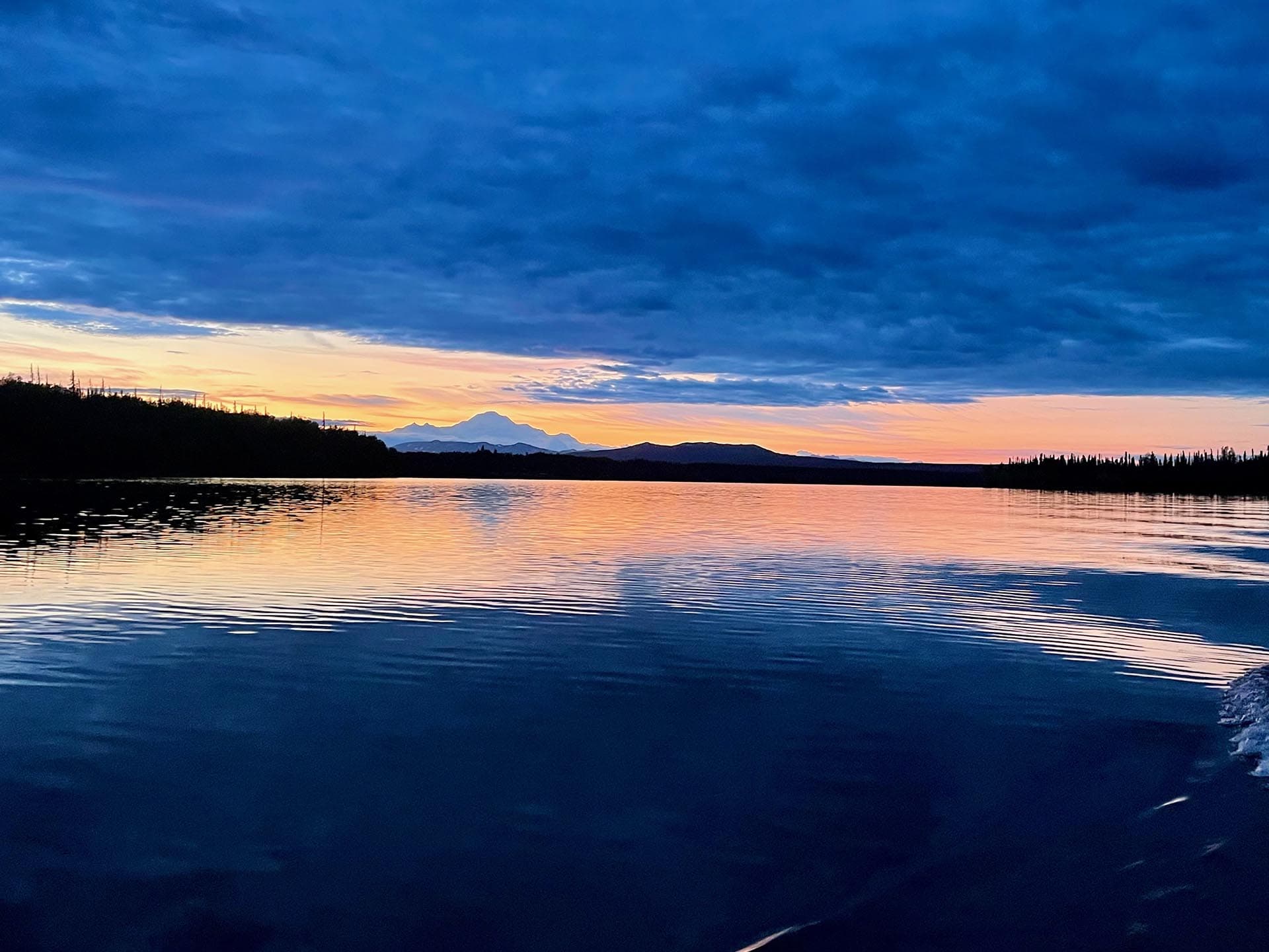 Silhouetted shoreline and distant mountains at sunset over a calm lake.