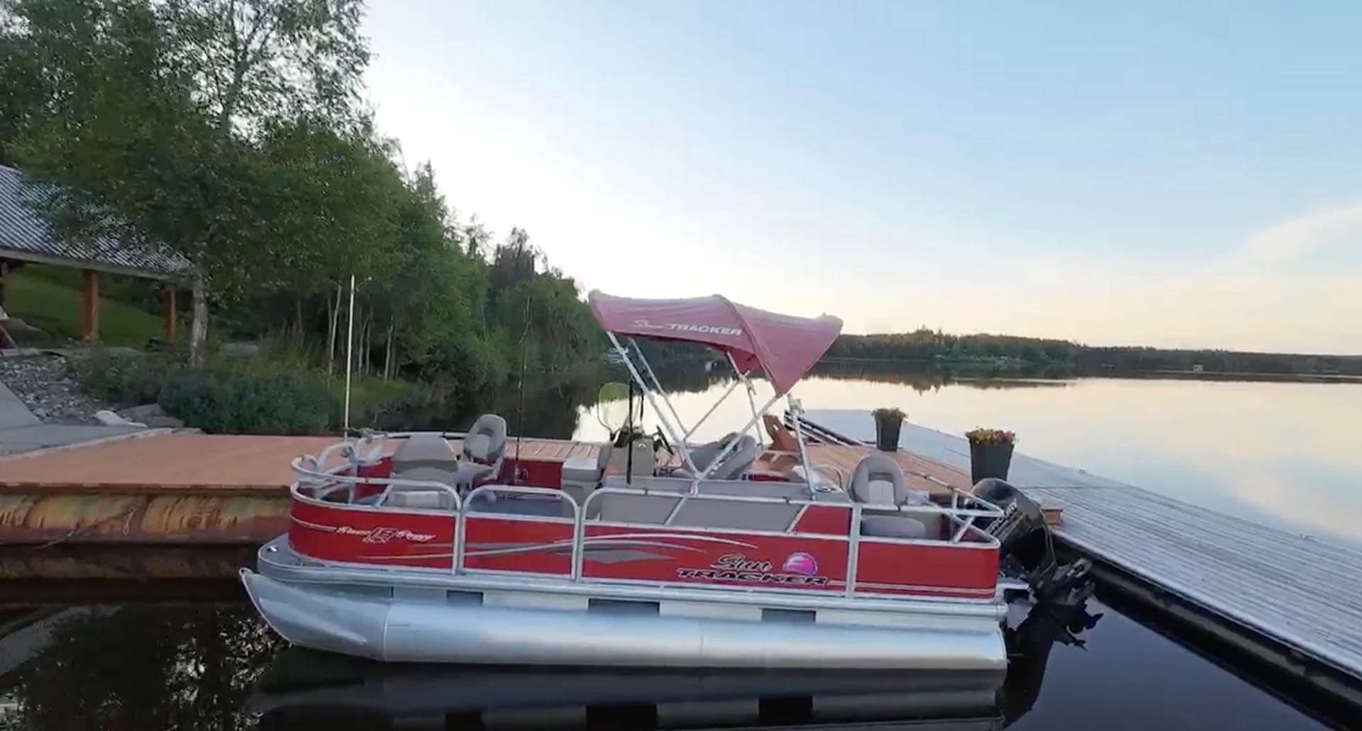 Pontoon boat with pink canopy docked at a calm lake, bordered by trees and a wooden pier on a clear day.
